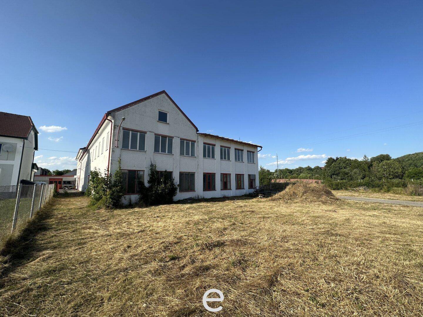 Älteres Wohn- oder Bürogebäude mit mehreren Fenstern, umgeben von trockenem Gras und Bäumen, mit Renovierungsbedarf.