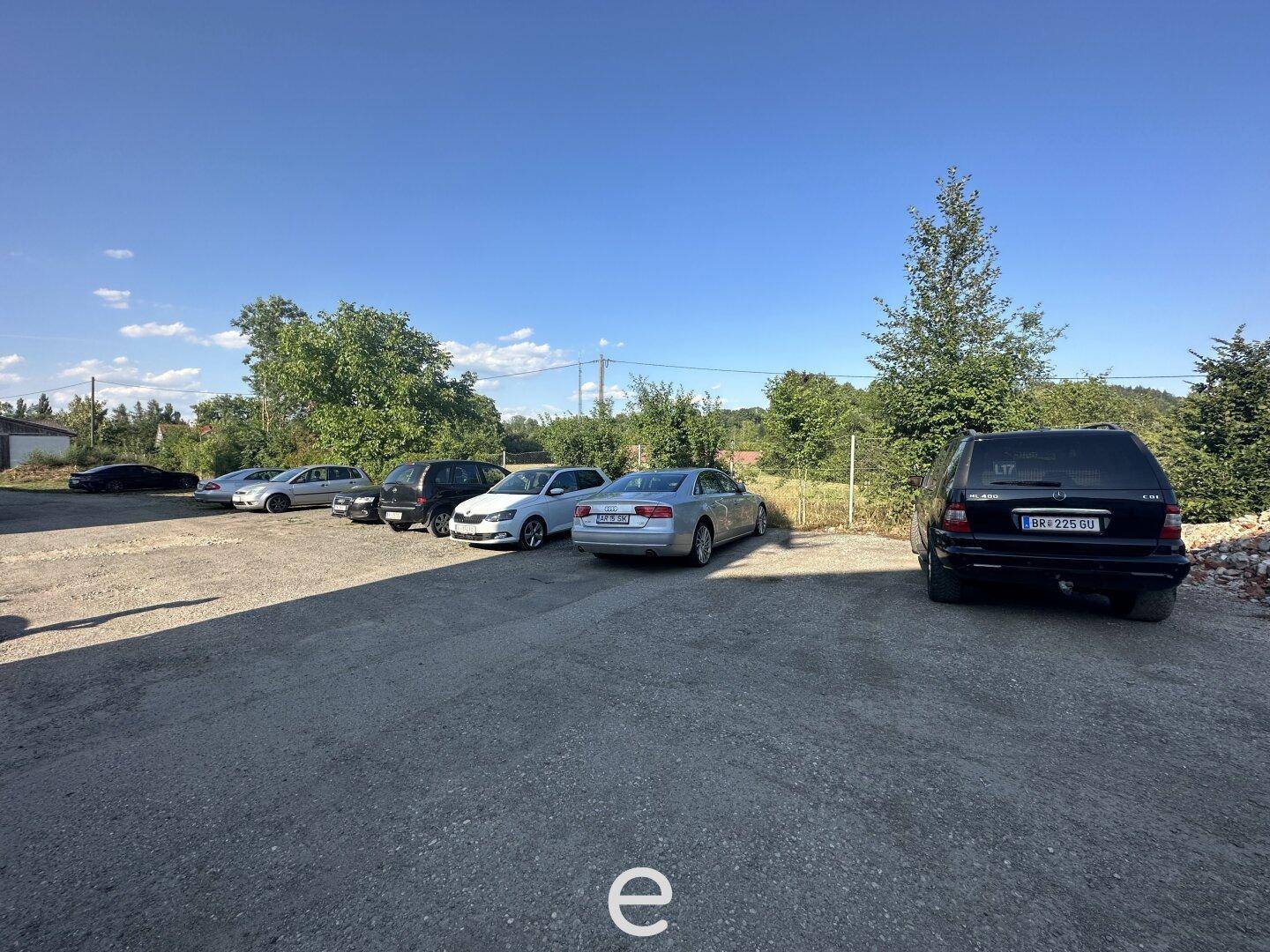 Großer Außenparkplatz mit mehreren geparkten Fahrzeugen, umgeben von Bäumen und Büschen unter blauem Himmel.