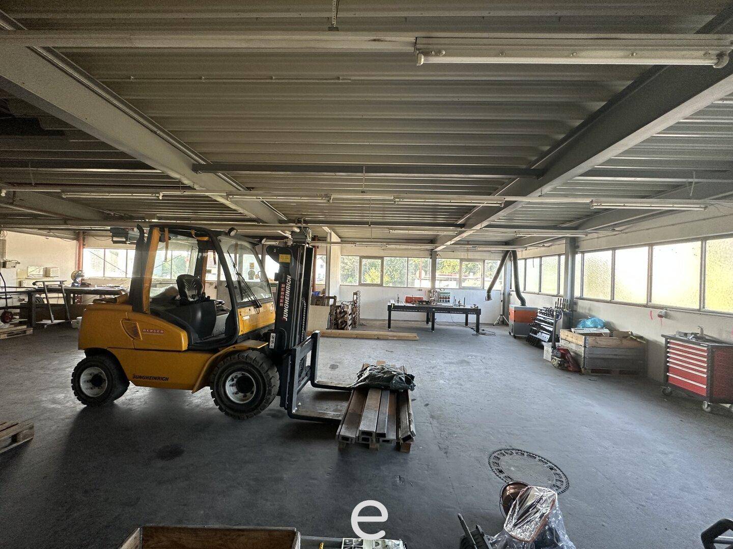Geräumige Lagerhalle mit einem Gabelstapler, großen Fenstern und sichtbaren Stahlträgern, die Renovierungsbedarf aufweist.