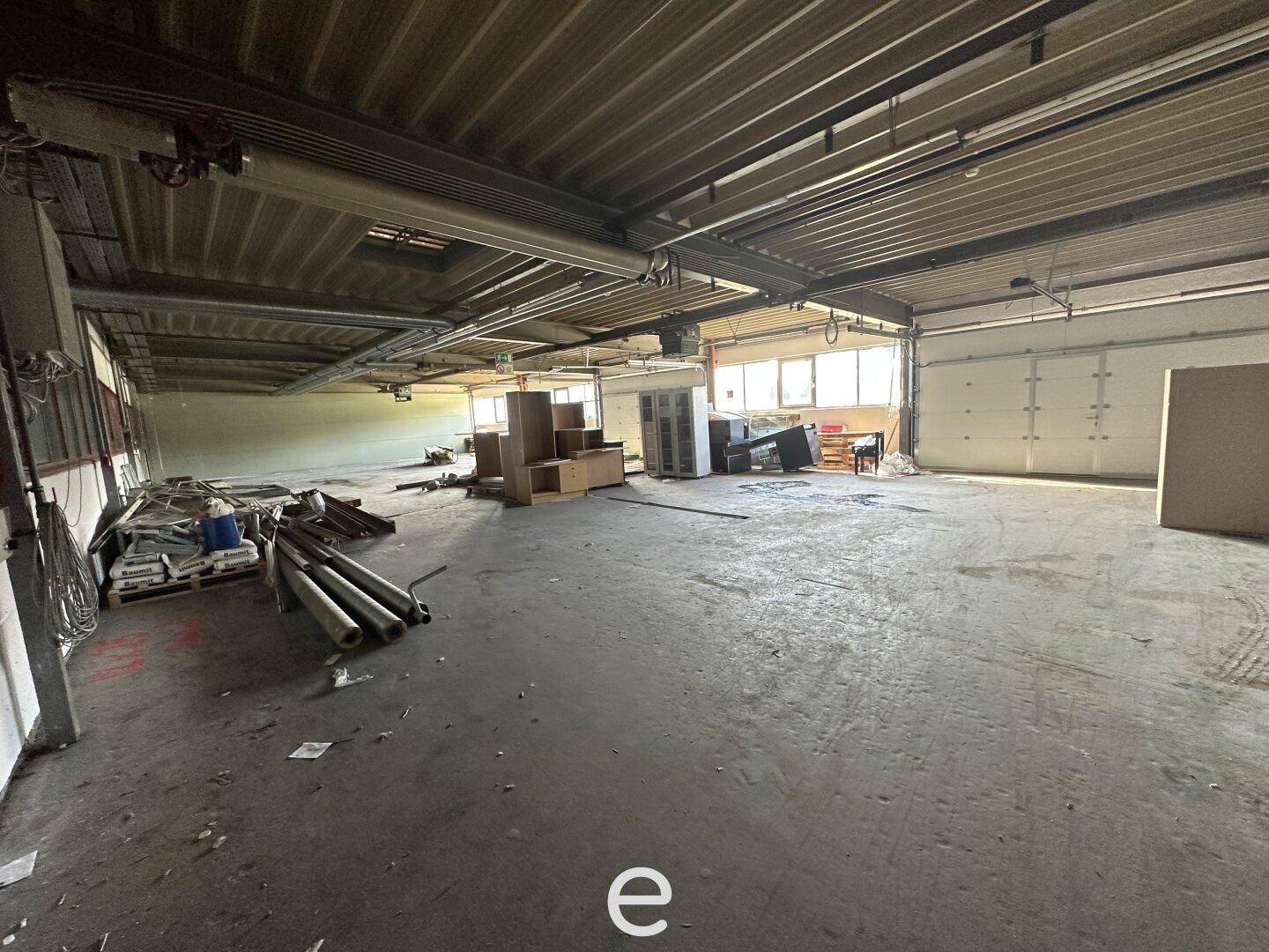 Weitläufige Lagerhalle mit Betonboden, Rolltoren und verschiedenen Gegenständen im Raum verteilt, mit Tageslicht.