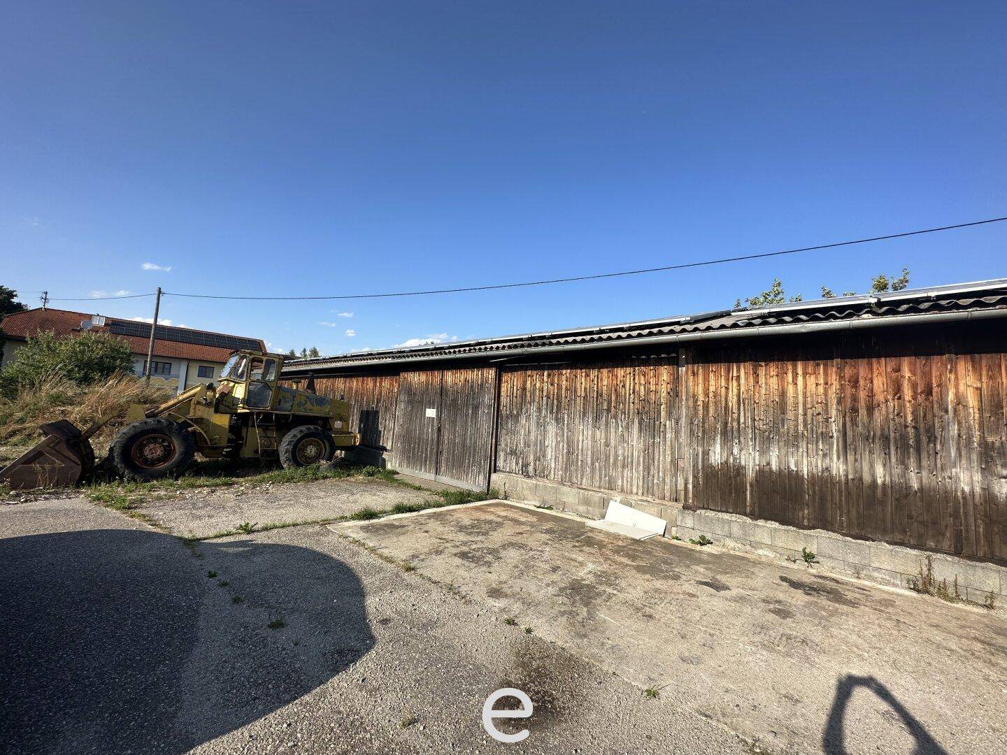 Altes Holzgebäude mit einer Baumaschine davor und einem Schotterplatz, das Renovierungsbedarf aufweist.