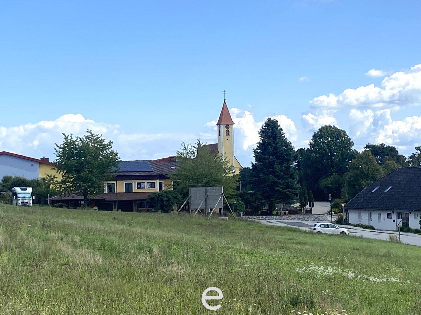 Ländliche Szene mit einer Kirche, Bäumen und weiteren Gebäuden unter einem klaren blauen Himmel.