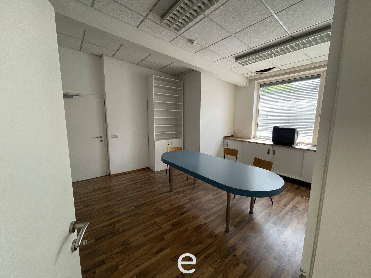 Funktionales Büro mit Holzboden, einem ovalen Tisch und Einbauschränken unter dem Fenster, ideal für Besprechungen.