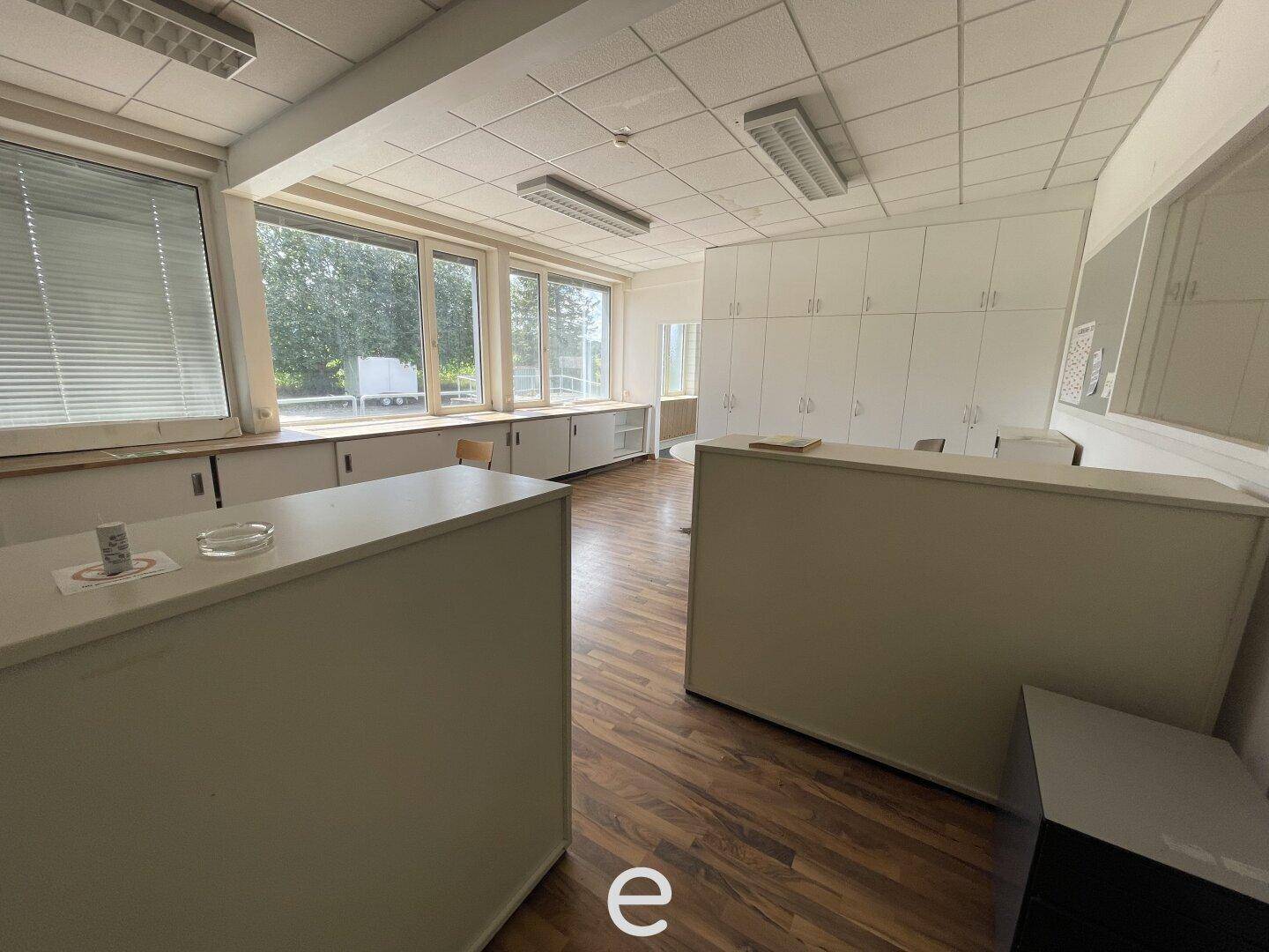 Helles Büro mit großen Fenstern, Holzboden und weißen Einbauschränken entlang der Wand, gut beleuchtet.