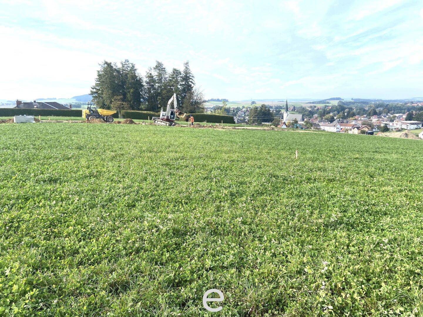 Weitläufiges grünes Feld mit Baumaschinen und malerischem Blick auf das umliegende Dorf und die Landschaft.