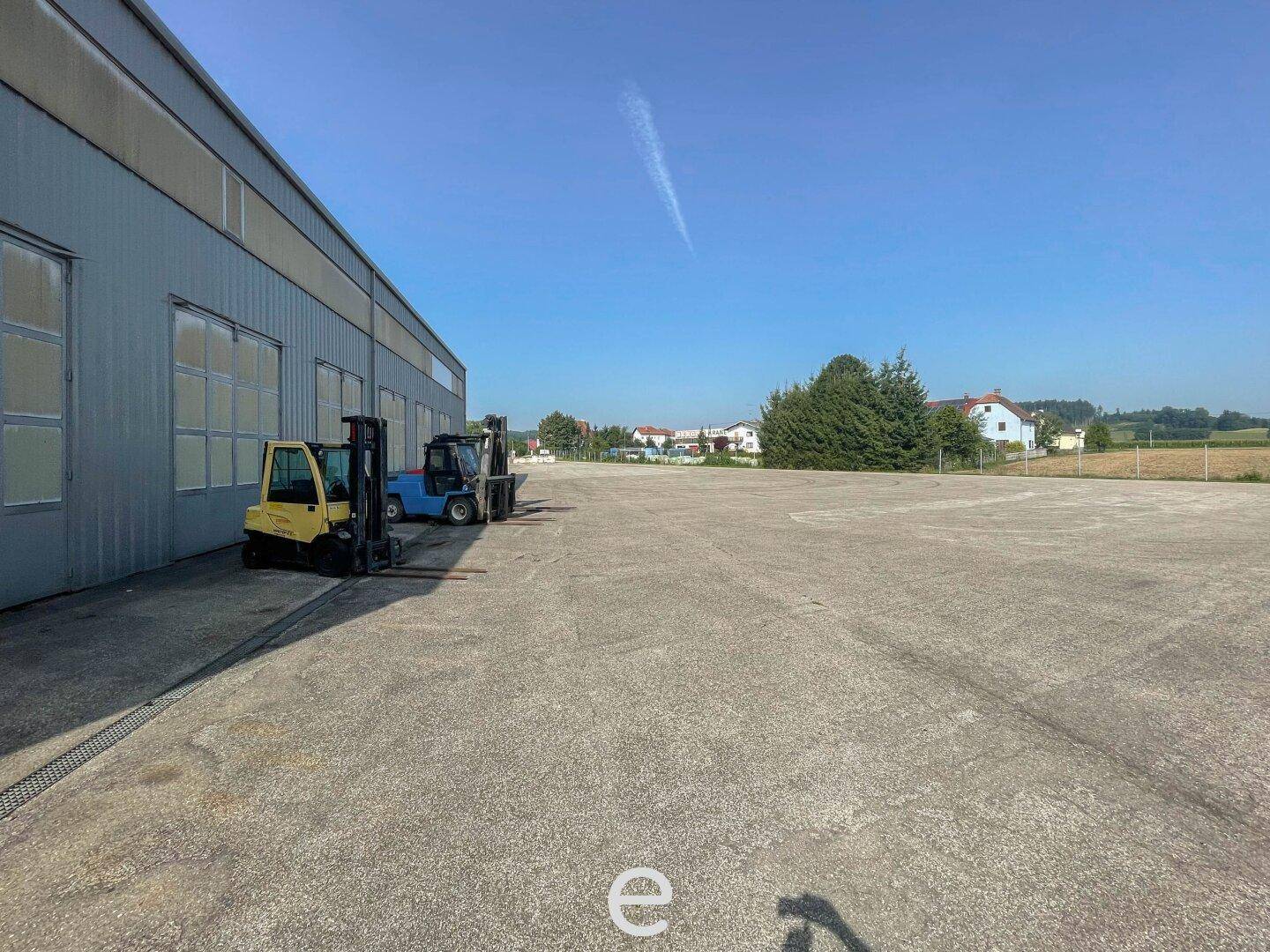 Außenansicht des Industriegebäudes mit großem, gepflastertem Vorplatz und geparkten Gabelstaplern unter blauem Himmel.
