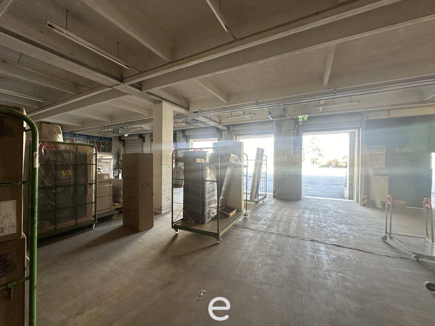 Lagerhalle mit Rollwagen und Kartons, Blick auf die hellen Laderampen und den Außenbereich.