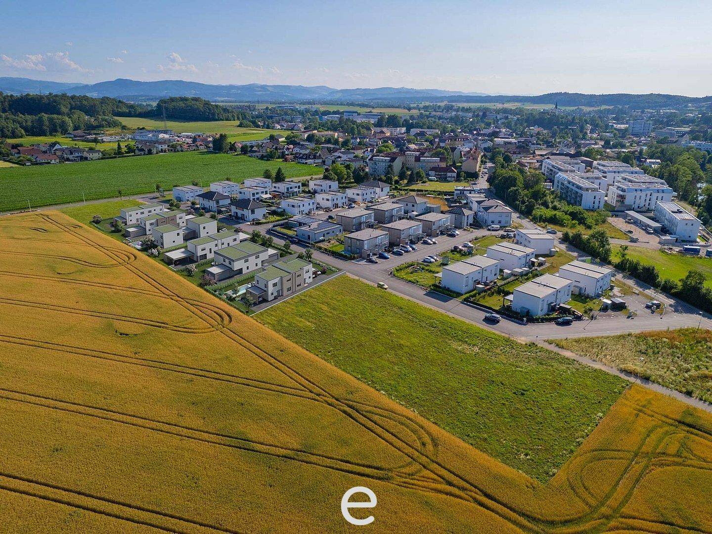 Luftaufnahme einer Wohnsiedlung umgeben von Feldern und grüner Landschaft unter blauem Himmel.