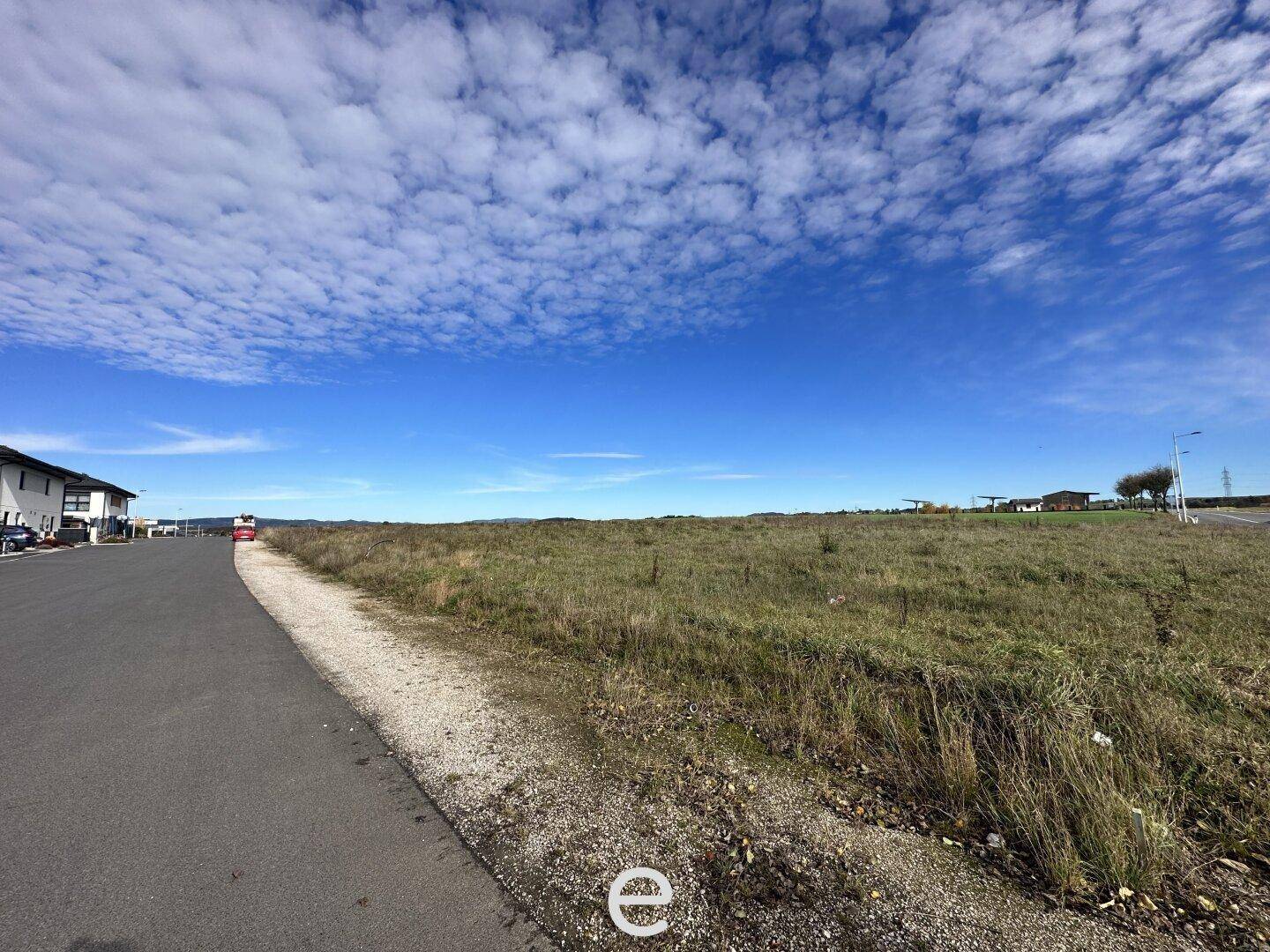 Blick über ein weites Feld mit Gras und Erde neben einer asphaltierten Straße unter blauem Himmel.