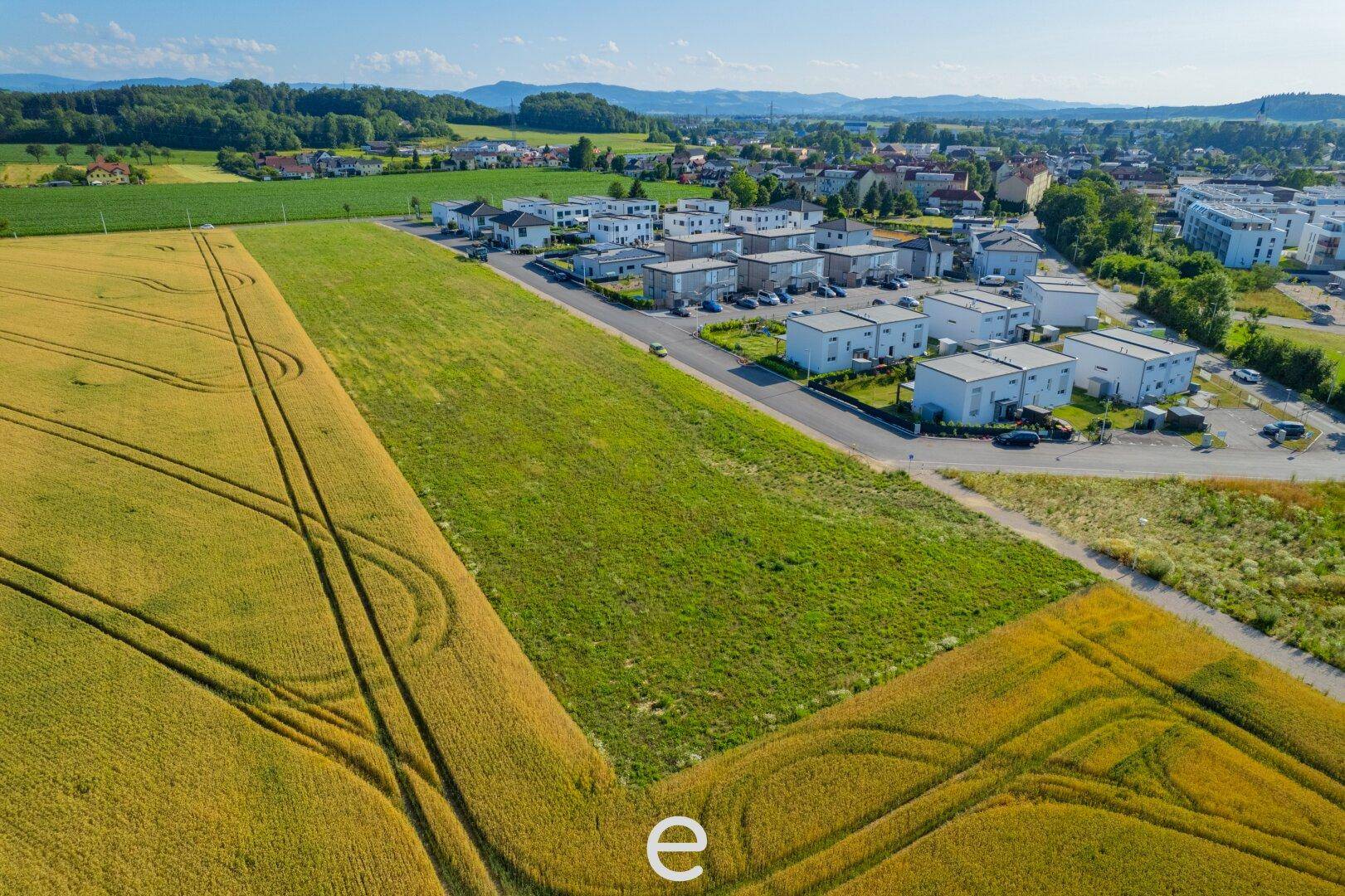 Luftaufnahme eines großen, grünen Baugrundstücks umgeben von Feldern und bestehenden Häusern.