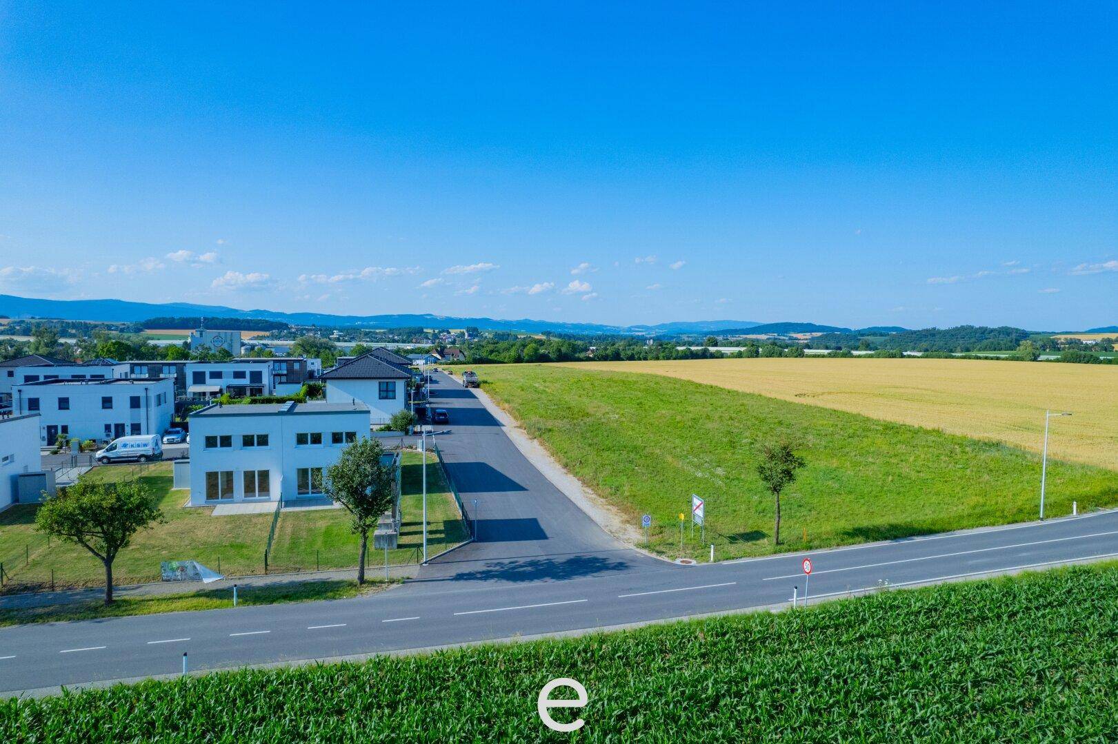 Luftaufnahme einer modernen Wohnsiedlung mit Straße und angrenzendem Feld unter blauem Himmel.