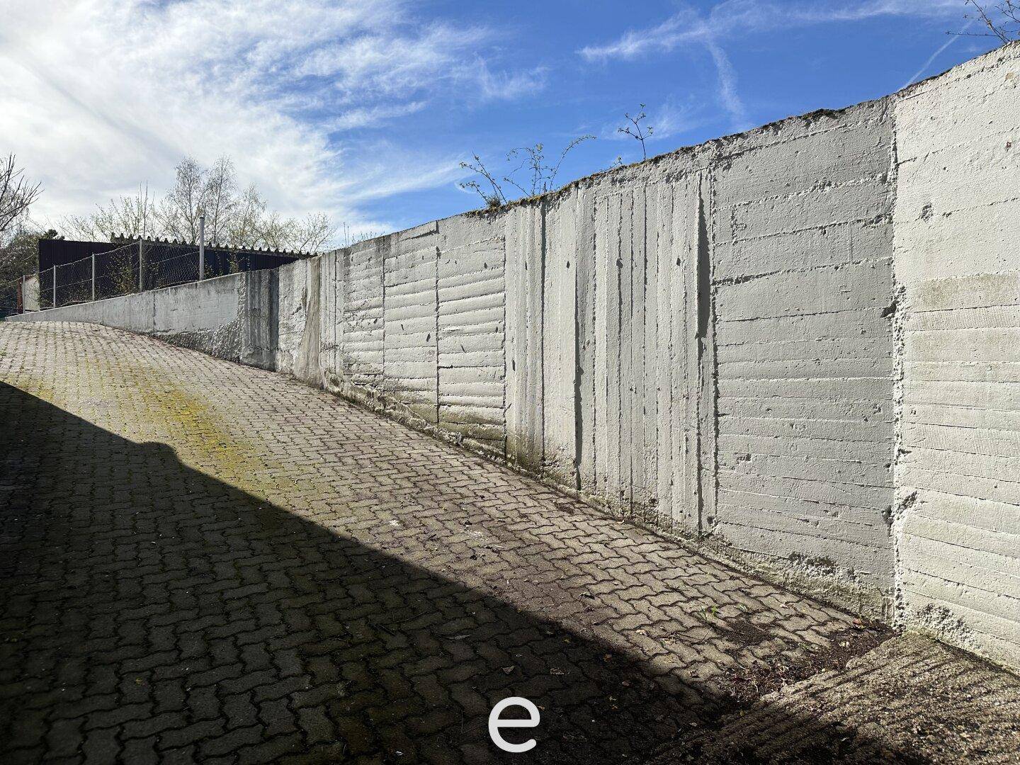 Eine gepflasterte Auffahrt führt entlang einer hohen Betonmauer unter blauem Himmel.