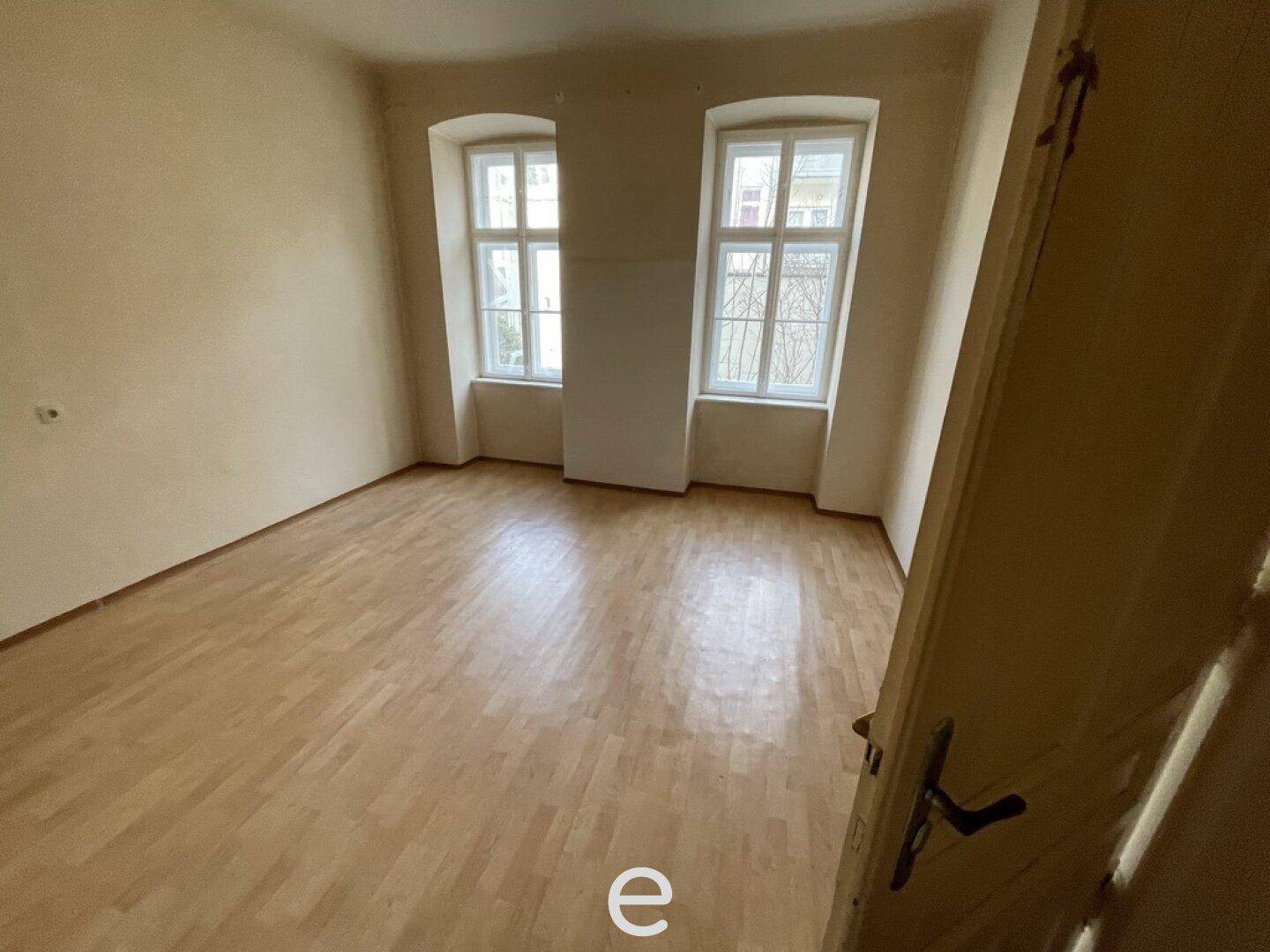 Heller, leerer Raum mit Holzboden und zwei großen Fenstern, die viel Tageslicht hereinlassen.