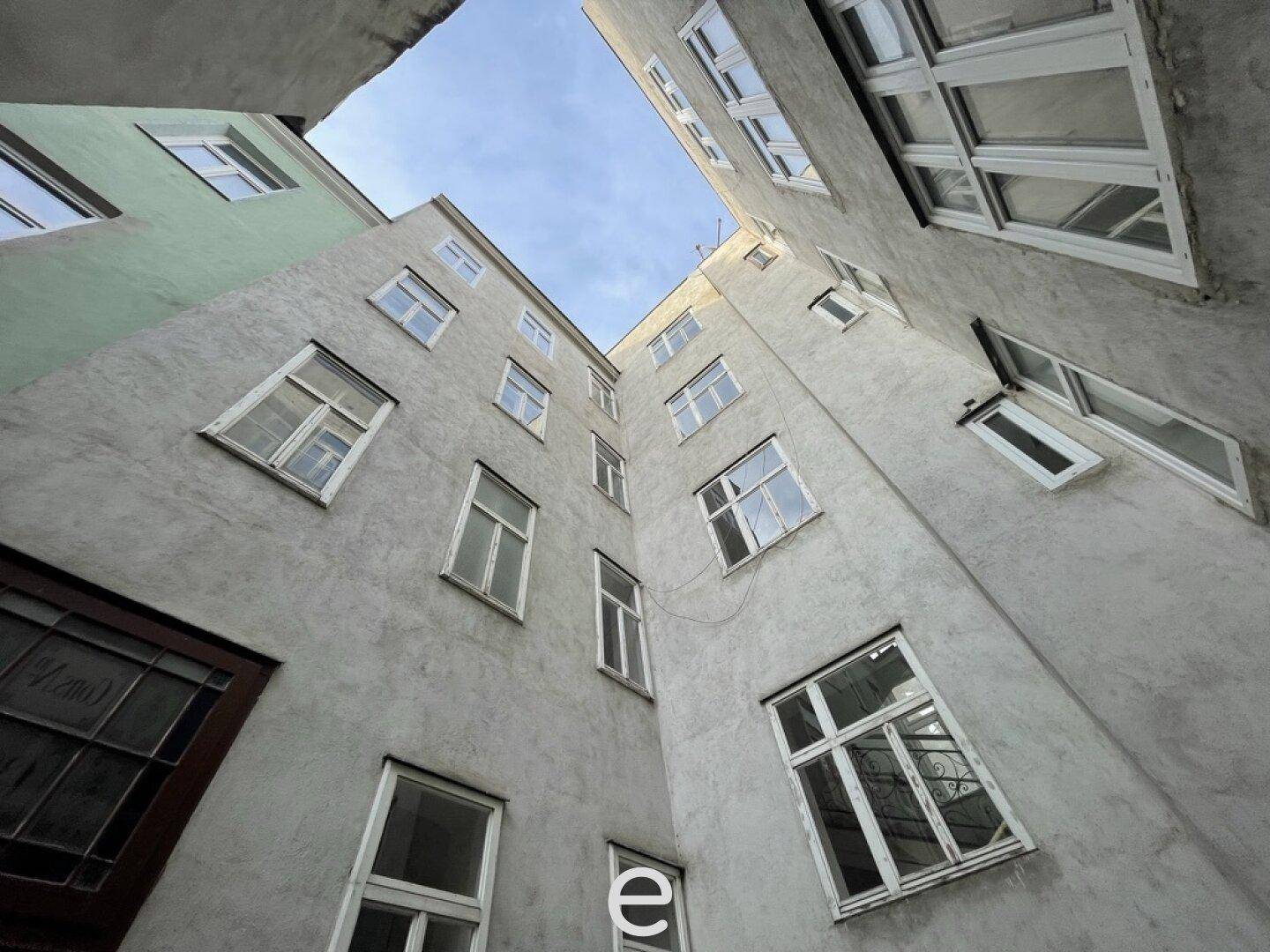 Blick nach oben in einen engen, grauen Innenhof mit zahlreichen Fenstern und klarem Himmel.