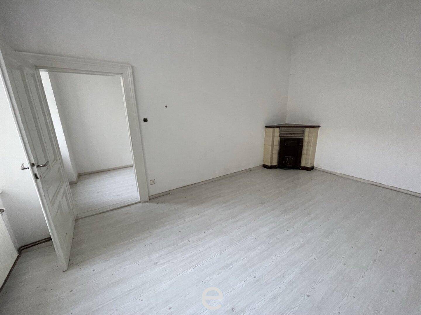 Leerer Raum mit hellem Boden, Zugang zu einem weiteren Zimmer und einem traditionellen Kachelofen.
