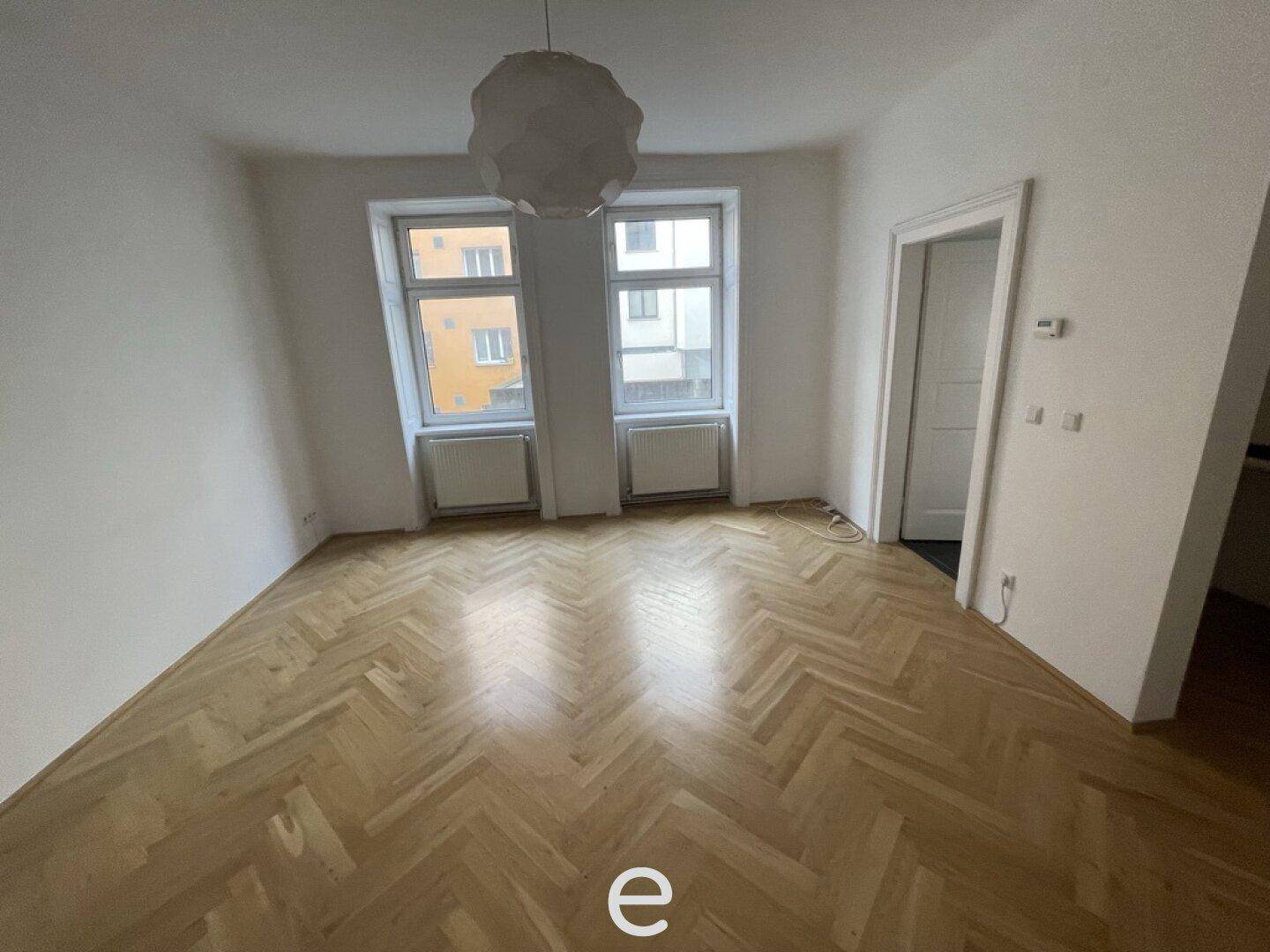 Geräumiges Wohnzimmer mit Fischgrätparkettboden und zwei großen Fenstern, die viel Licht hereinlassen.