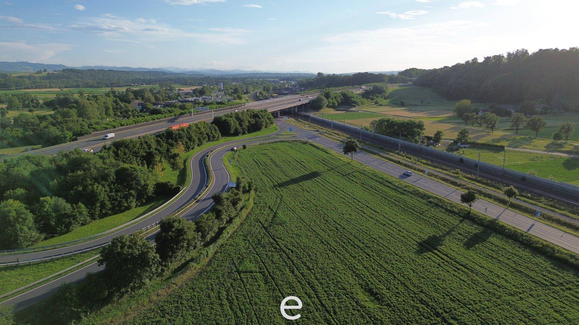 Weitläufige Luftaufnahme einer Autobahn mit angrenzenden Feldern und bewaldeten Hügeln unter sonnigem Himmel.