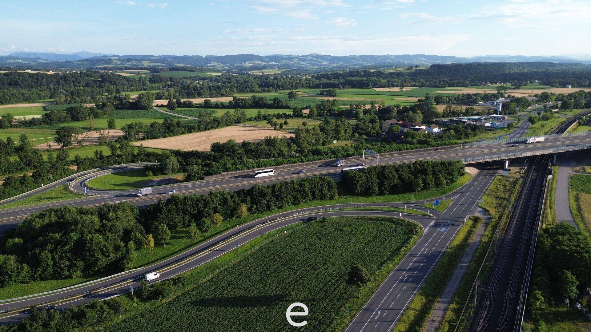 Panoramablick aus der Luft auf eine belebte Autobahnkreuzung, umgeben von weiten Feldern und sanften Hügeln.