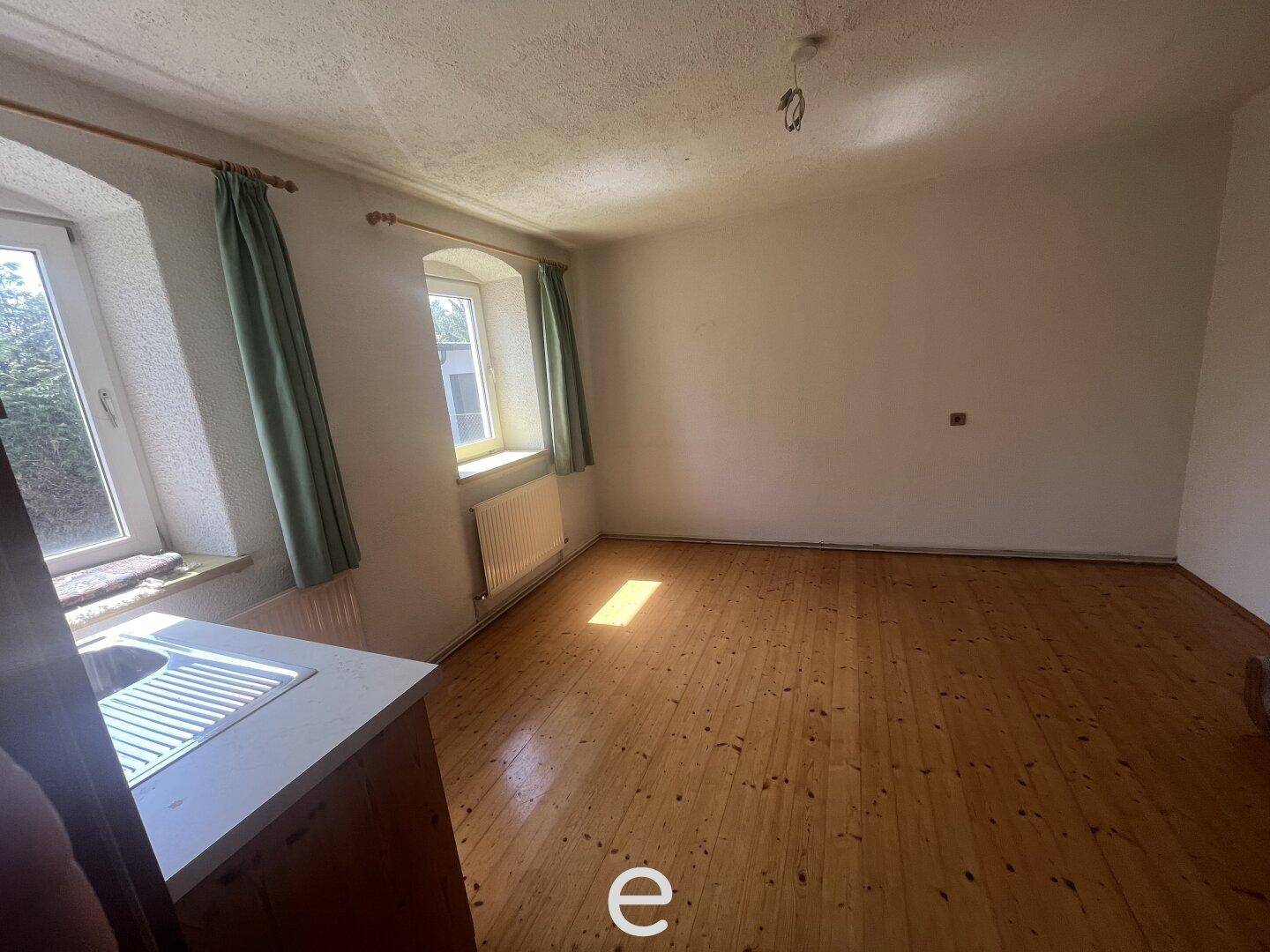 Leerer Raum mit Holzboden, zwei Fenstern und einem Heizkörper unter der Fensterbank.
