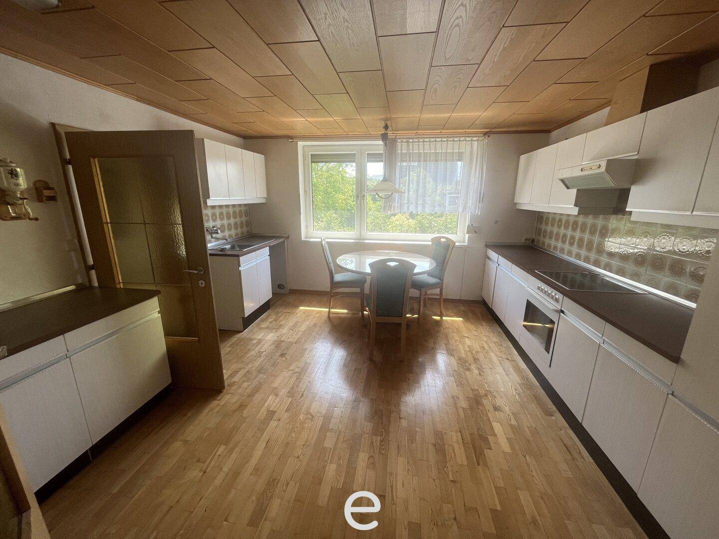 Küche mit Holzboden, weißen Schränken, Esstisch und Blick ins Grüne durch das Fenster.