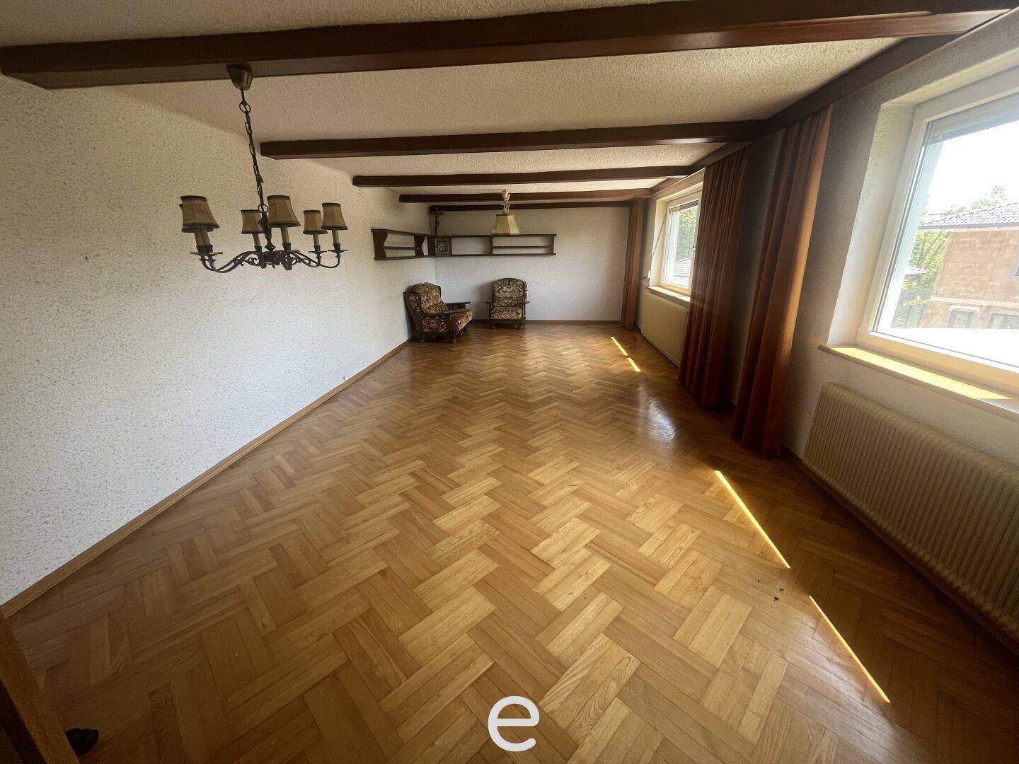 Wohnzimmer mit Fischgrätparkett, Holzbalken an der Decke und großen Fenstern.
