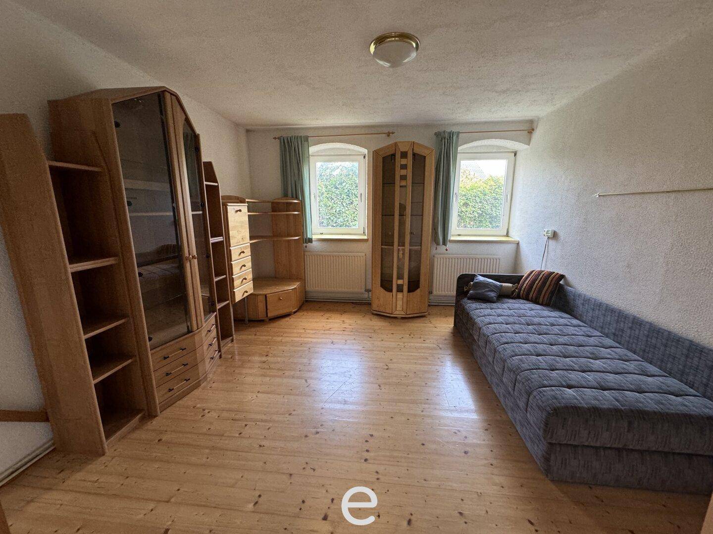 Schlafzimmer mit Holzboden, zwei Fenstern und einem Sofa sowie Holzregalen.