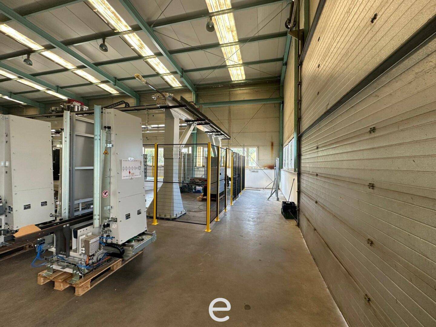 Blick in die Industriehalle mit großen Maschinen, gelben Schutzgittern und Fenstern, die Tageslicht hereinlassen.