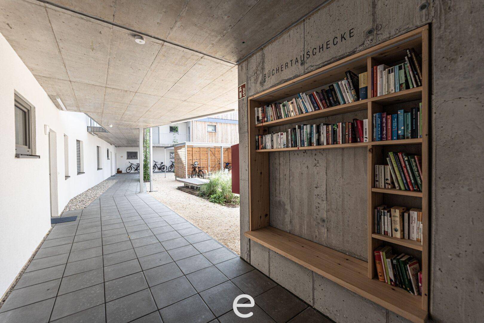 Überdachter Gang mit Betonwänden und einem integrierten Bücherregal als Büchertauschbörse für die Bewohner.