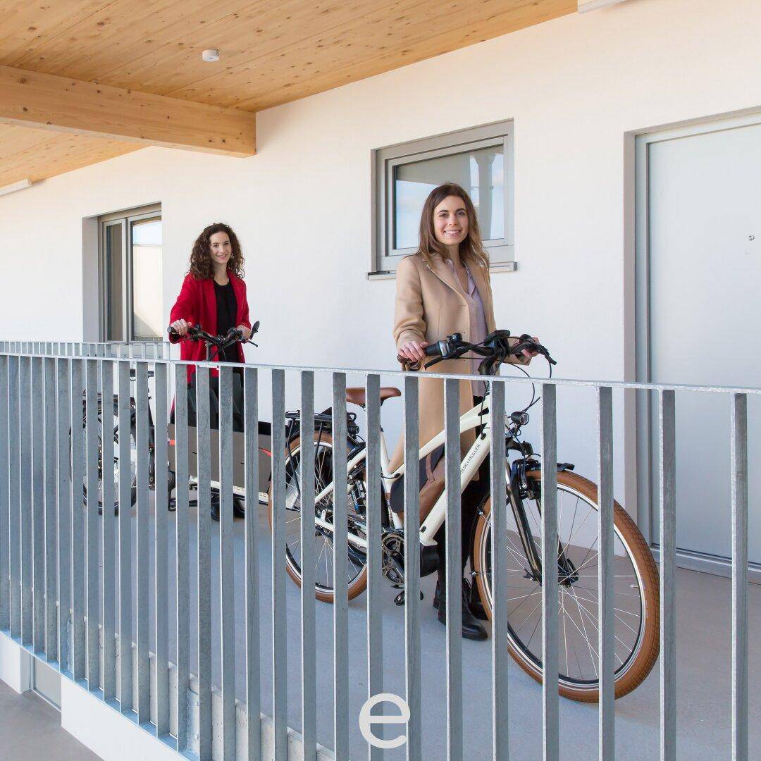 Zwei Frauen mit Fahrrädern in einem hellen, überdachten Gemeinschaftsbereich mit moderner Architektur und Holzdecke.