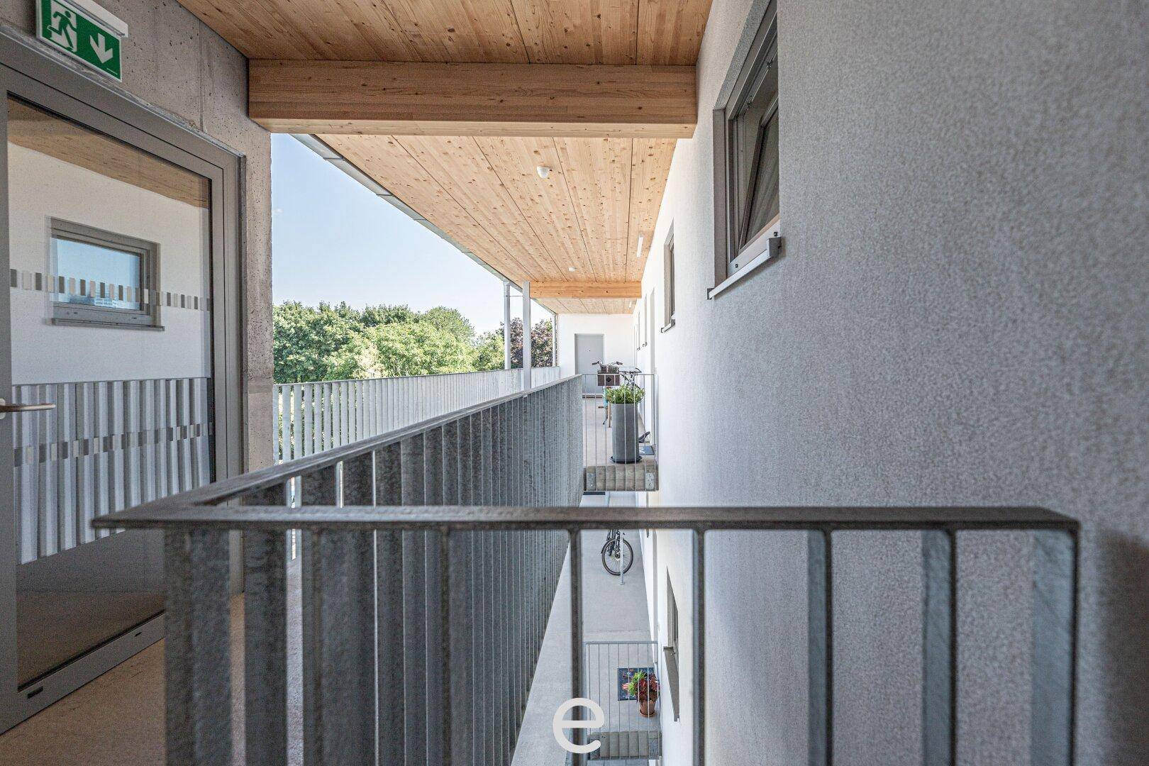 Außenflur mit Holzdecke und Metallgeländer, bietet einen Blick auf den Innenhof der Wohnanlage.