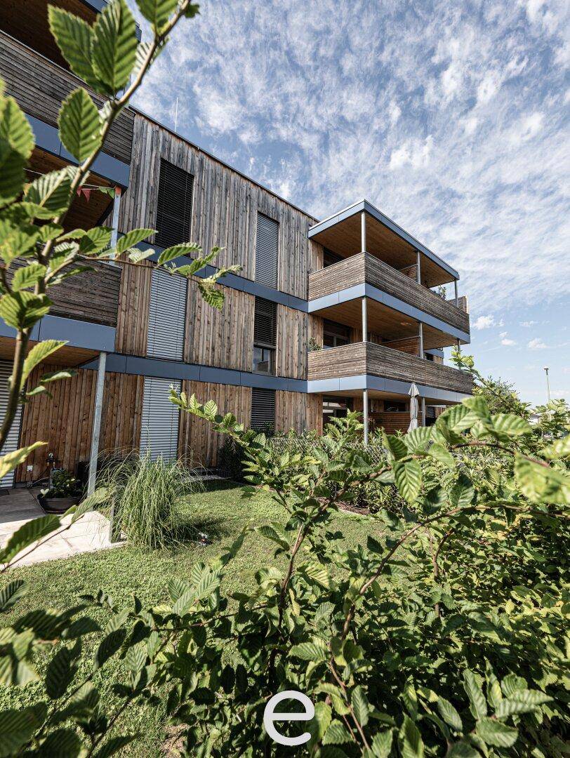 Moderne Holzfassade eines Wohngebäudes, umgeben von üppiger Vegetation unter blauem Himmel.