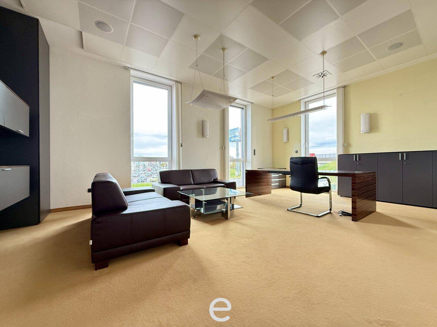 Großes Büro mit Teppichboden, bequemen Sofas und zwei Arbeitsplätzen mit Blick nach draußen.