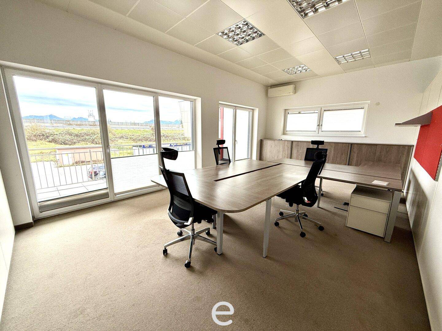 Helles Büro mit Teppichboden, großem Konferenztisch und Zugang zu einem Balkon.
