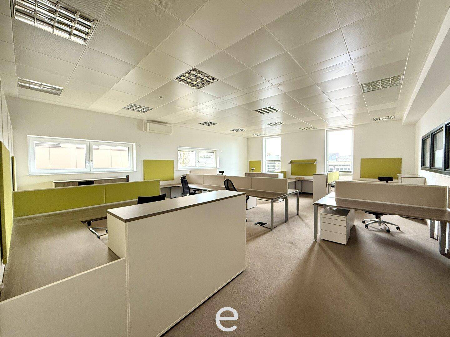 Großraumbüro mit mehreren Arbeitsplätzen, hellen Trennwänden und großen Fenstern für viel Tageslicht.