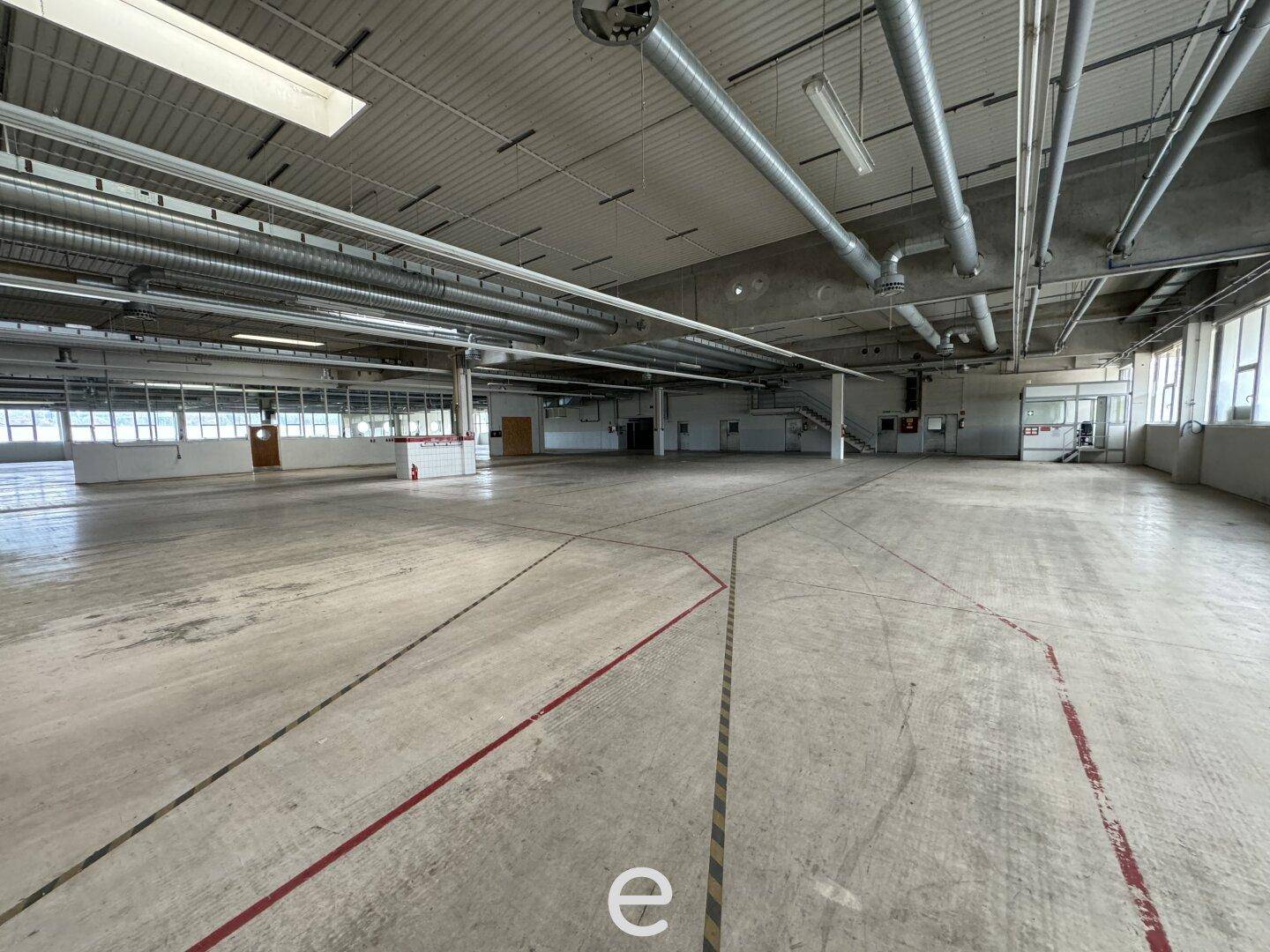 Große, leere Lagerhalle mit Betonboden, hohen Decken und sichtbaren Lüftungsrohren und Leitungen.