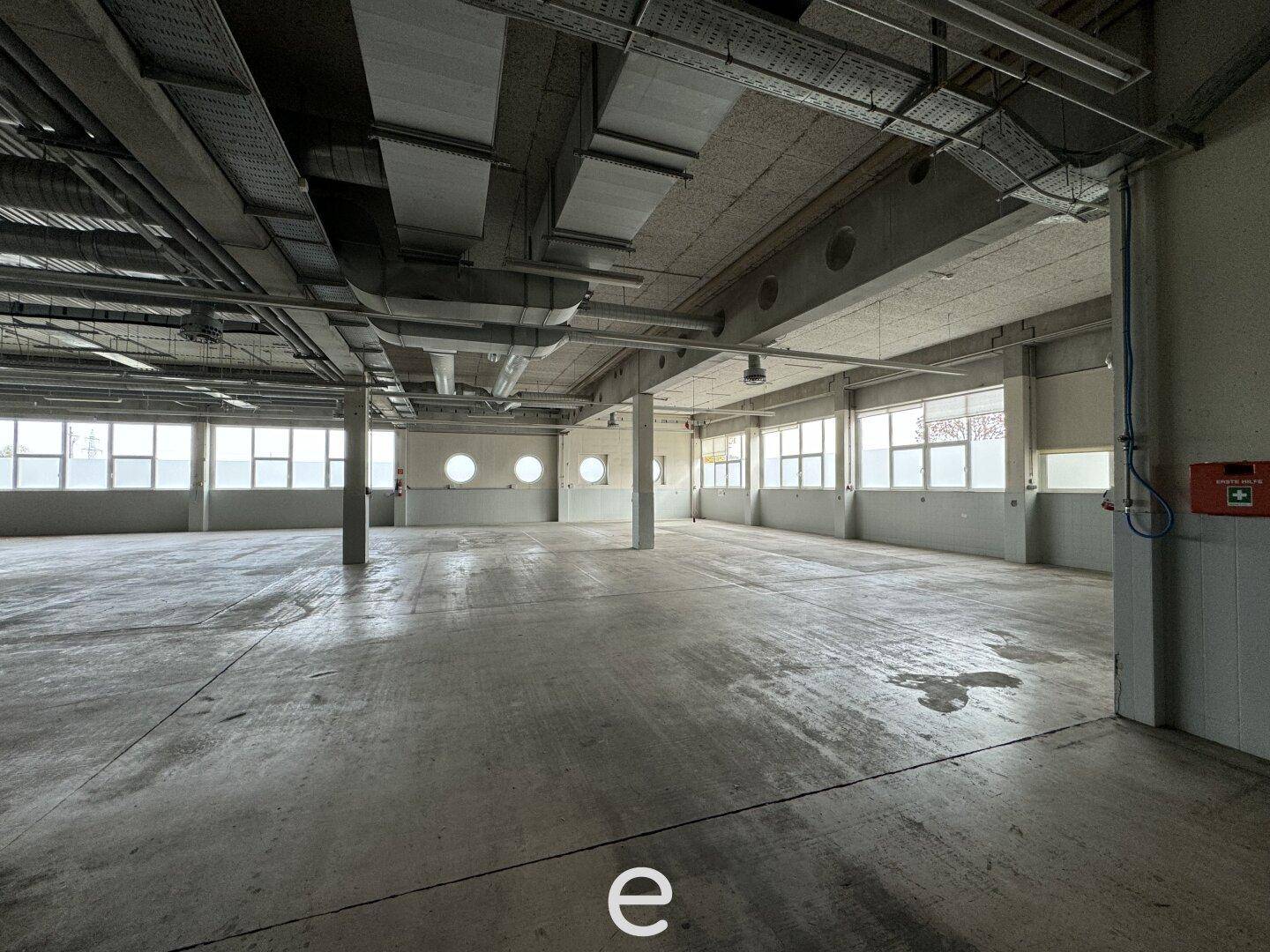 Weitläufige Halle mit Betonboden, vielen Fenstern und sichtbaren Deckeninstallationen und Rohren.