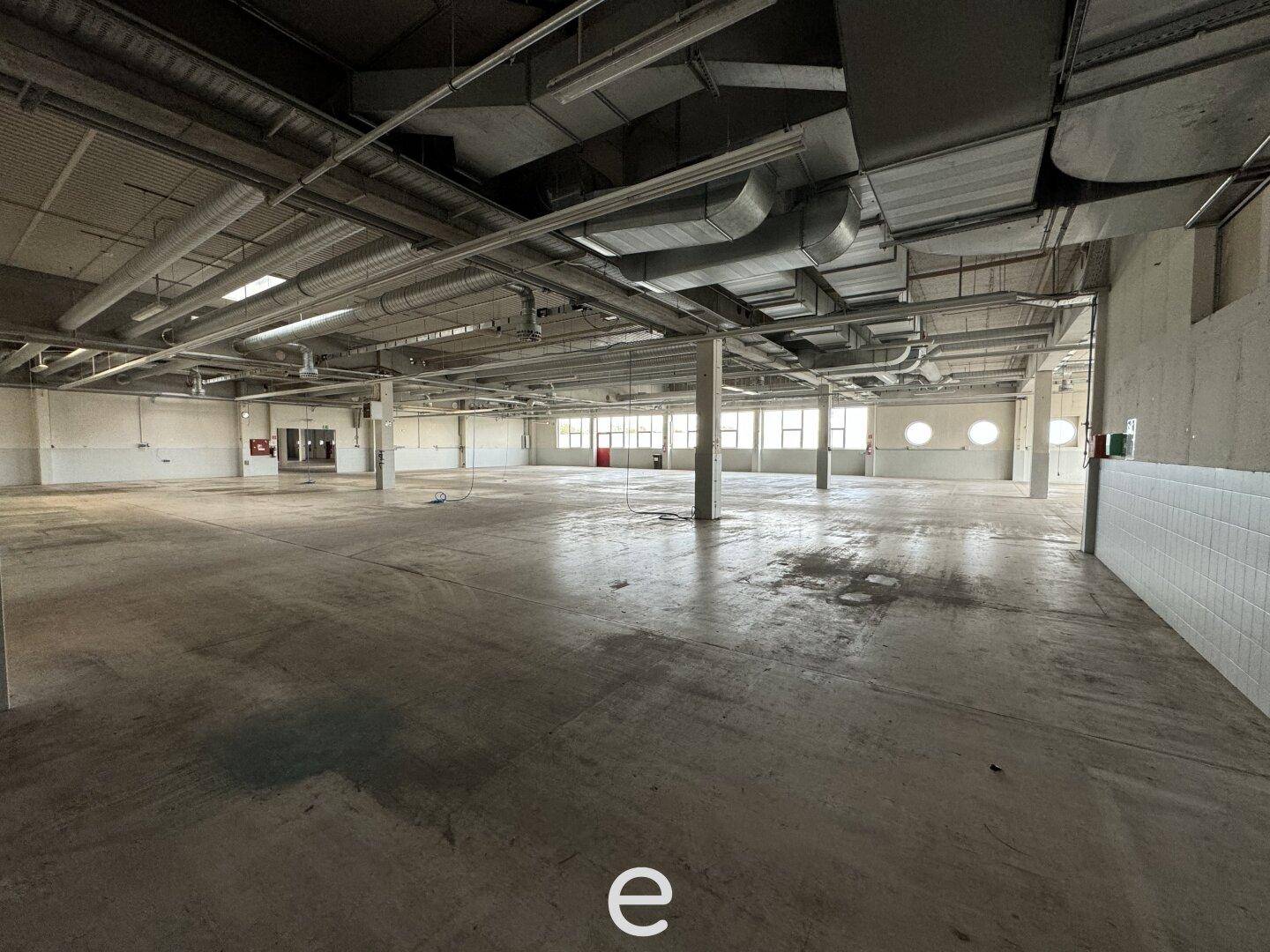 Große, leere Lagerhalle mit Betonboden, hohen Decken und sichtbaren Lüftungsrohren und Leitungen.
