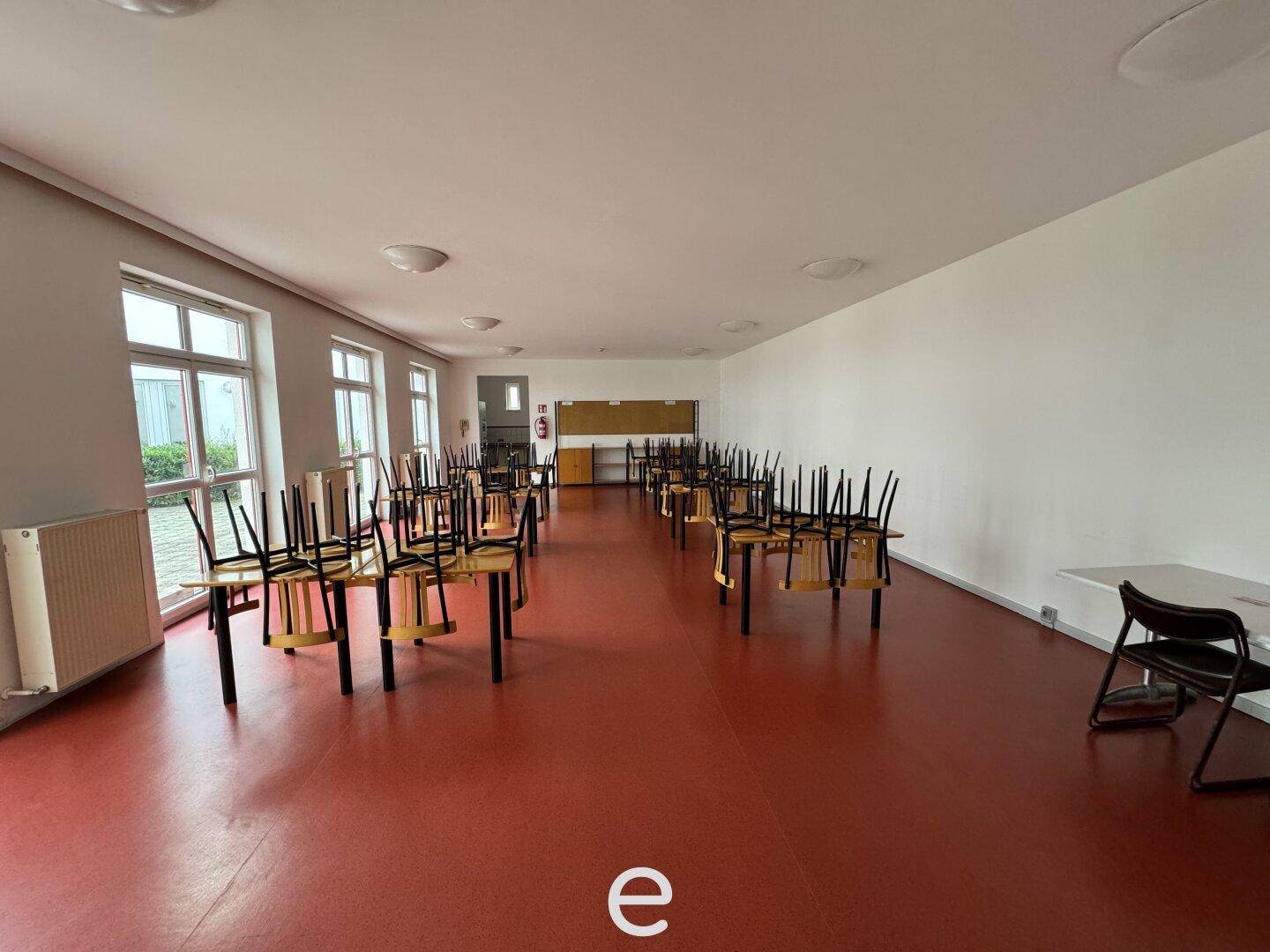 Kantine oder Mehrzweckraum mit rotem Boden, Tischen und gestapelten Stühlen sowie großen Fenstern.