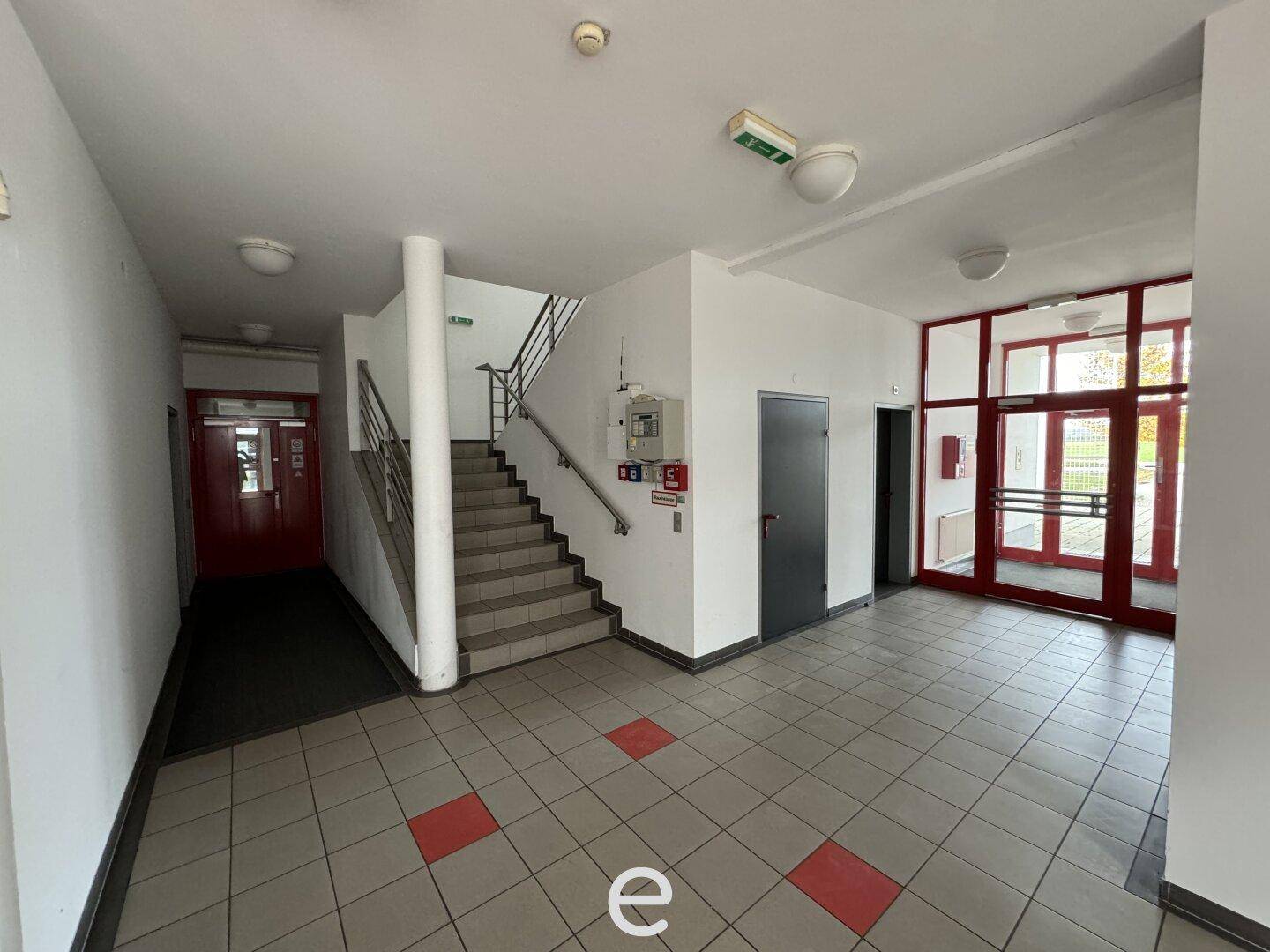 Heller Eingangsbereich mit Treppenhaus, Fliesenboden und großen Glastüren zum Außenbereich.