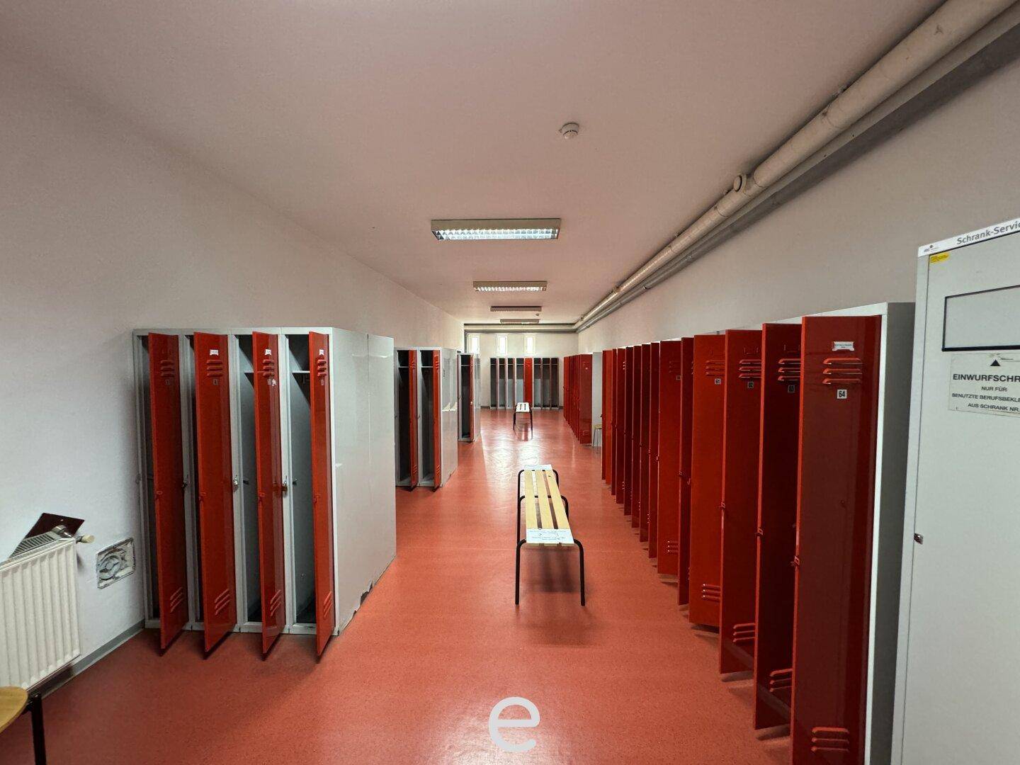 Umkleideraum mit Reihen von roten Spinden, einer Sitzbank und rotem Boden.