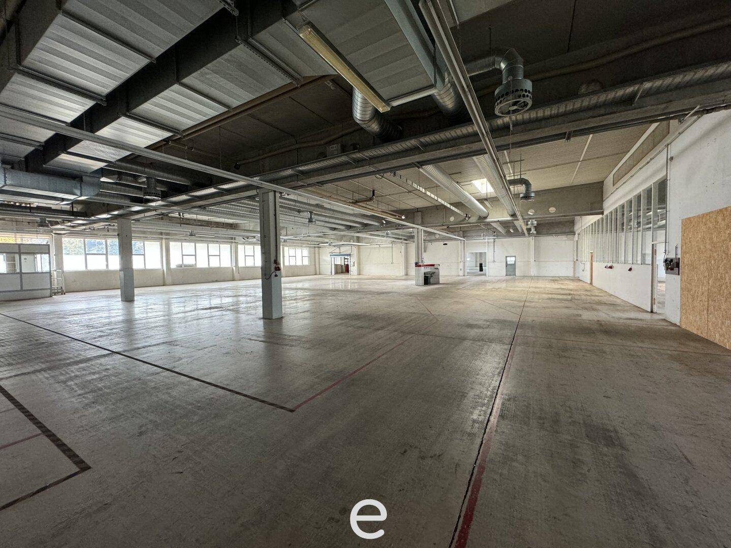 Weitläufige Lagerhalle mit Betonboden, hohen Decken und sichtbaren Lüftungsrohren und Leitungen.