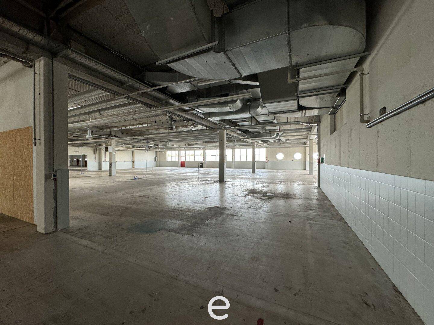 Geräumige Halle mit Betonboden, hohen Decken und sichtbaren Deckeninstallationen und Rohren.