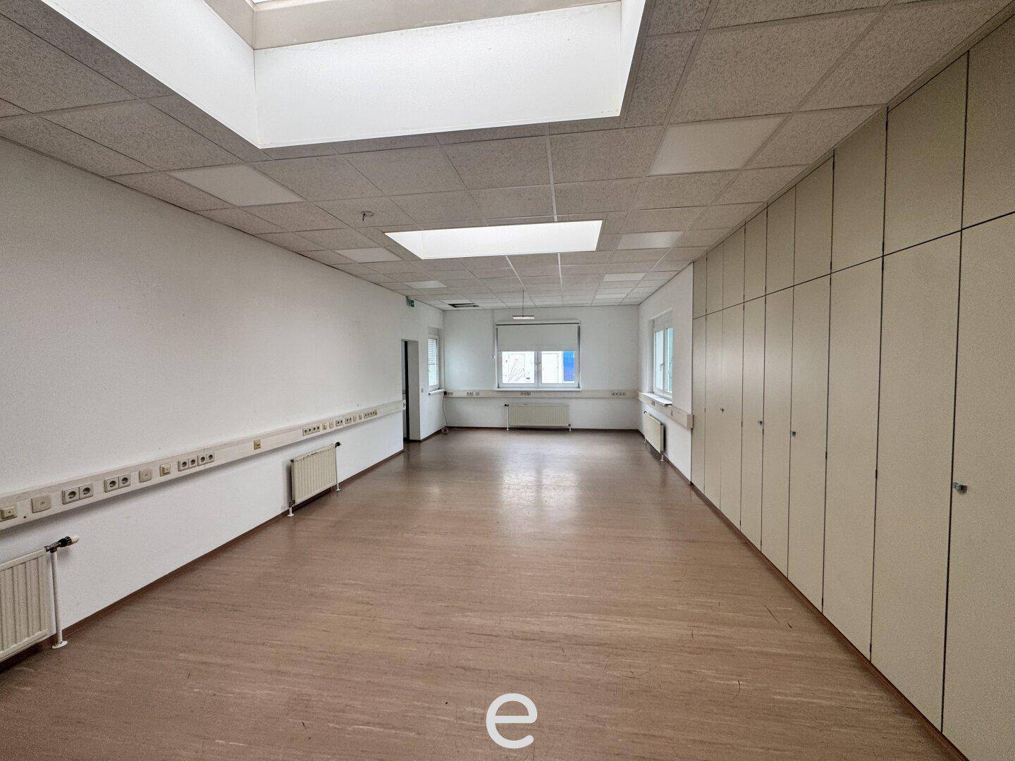 Langer Büroraum mit Einbauschränken, Fenstern und abgehängter Decke, gut beleuchtet.