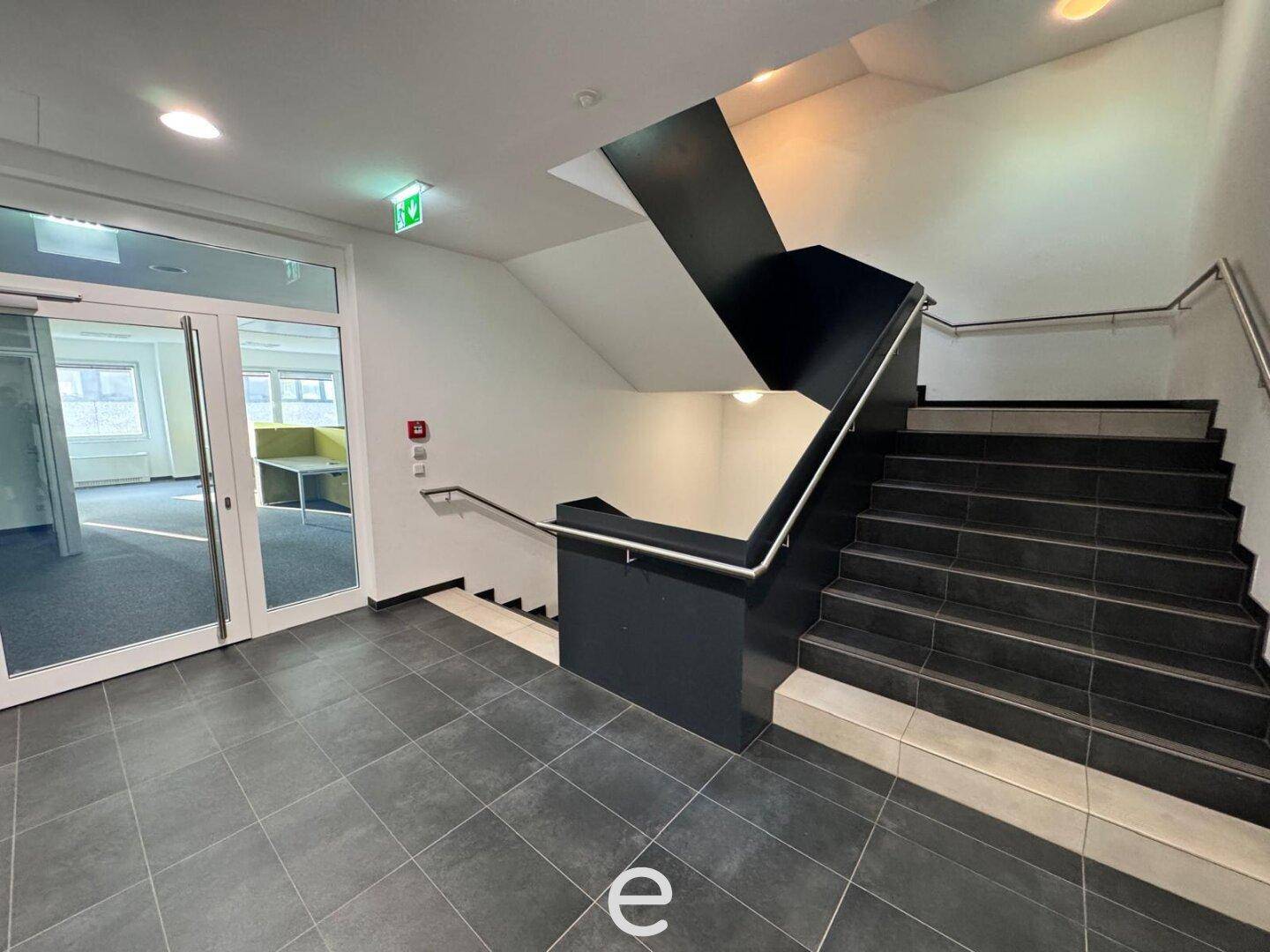 Modernes Treppenhaus mit dunklen Fliesenstufen und Geländer, das verschiedene Etagen verbindet, mit Zugang zu Büroräumen.
