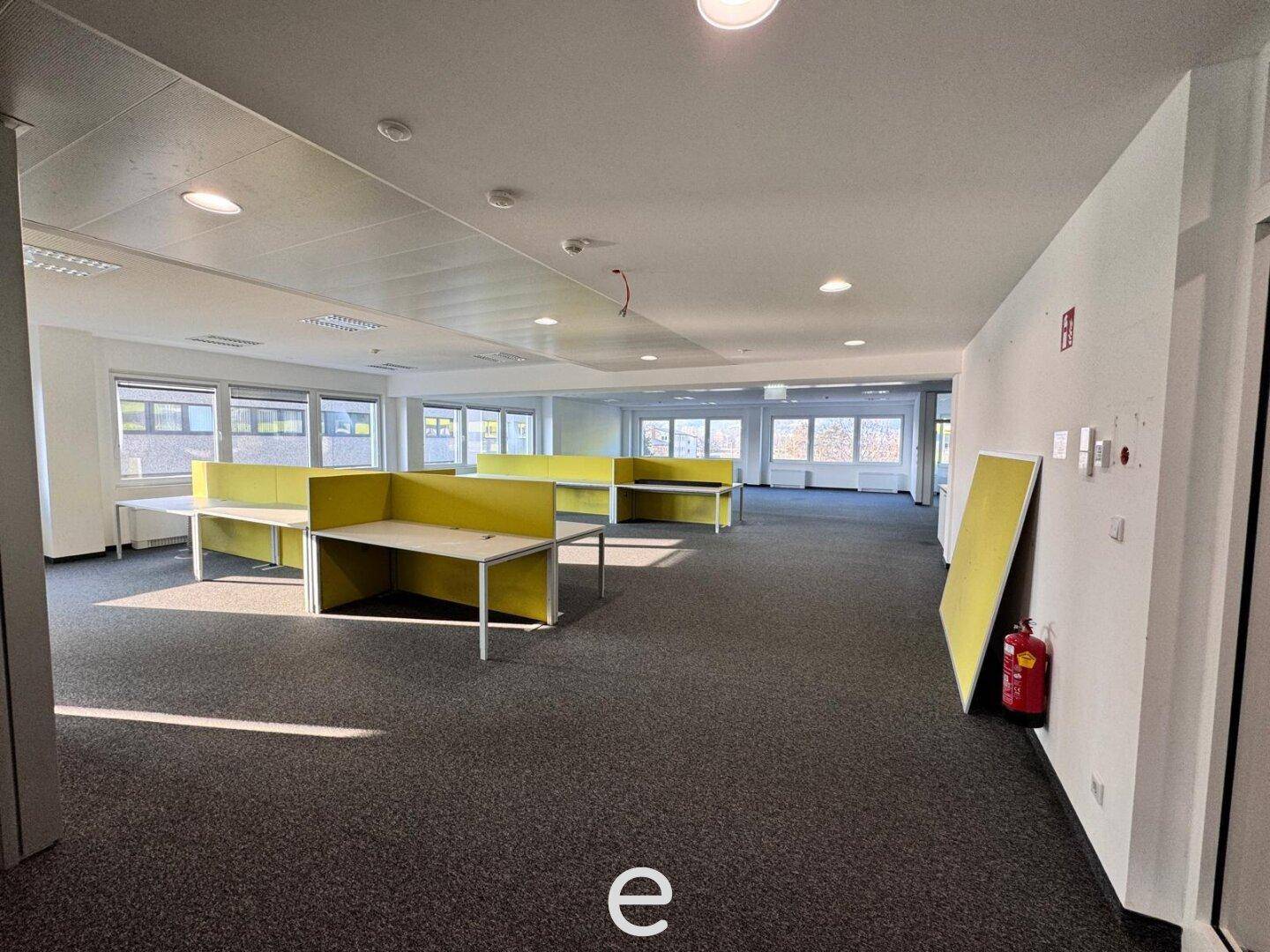 Weitläufiger Bürobereich mit zahlreichen Arbeitsplätzen und großen Fenstern, die viel Tageslicht hereinlassen, mit Teppichboden und Brandschutz.