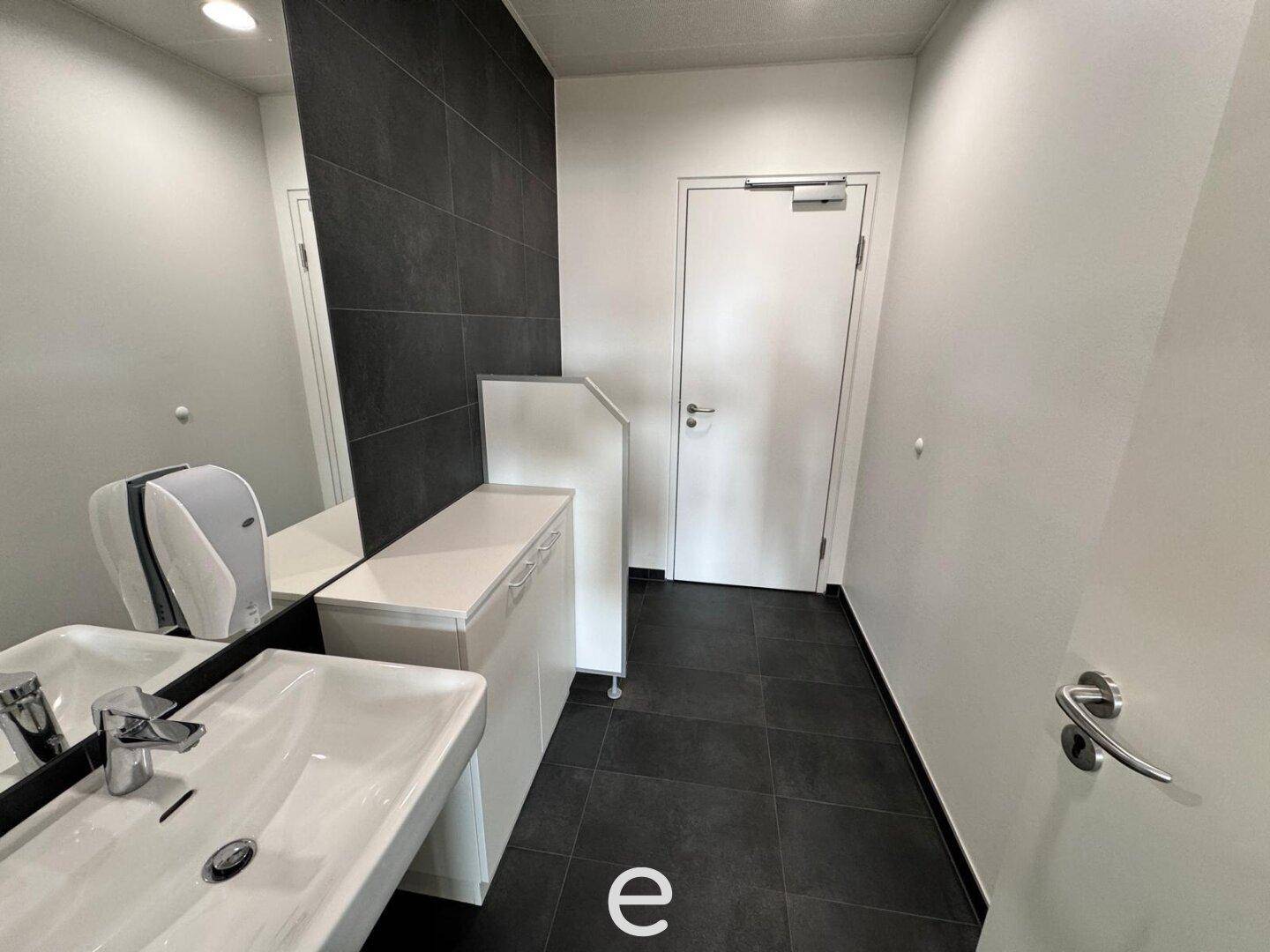 Modernes Badezimmer mit Waschbecken, großem Spiegel und dunklen Fliesen an Wand und Boden, in schlichtem und funktionalem Design.
