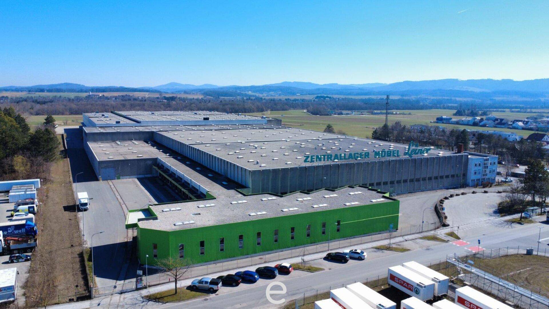 Luftaufnahme eines großen Industriegebäudes mit grüner Fassade und umliegenden Parkplätzen unter blauem Himmel.