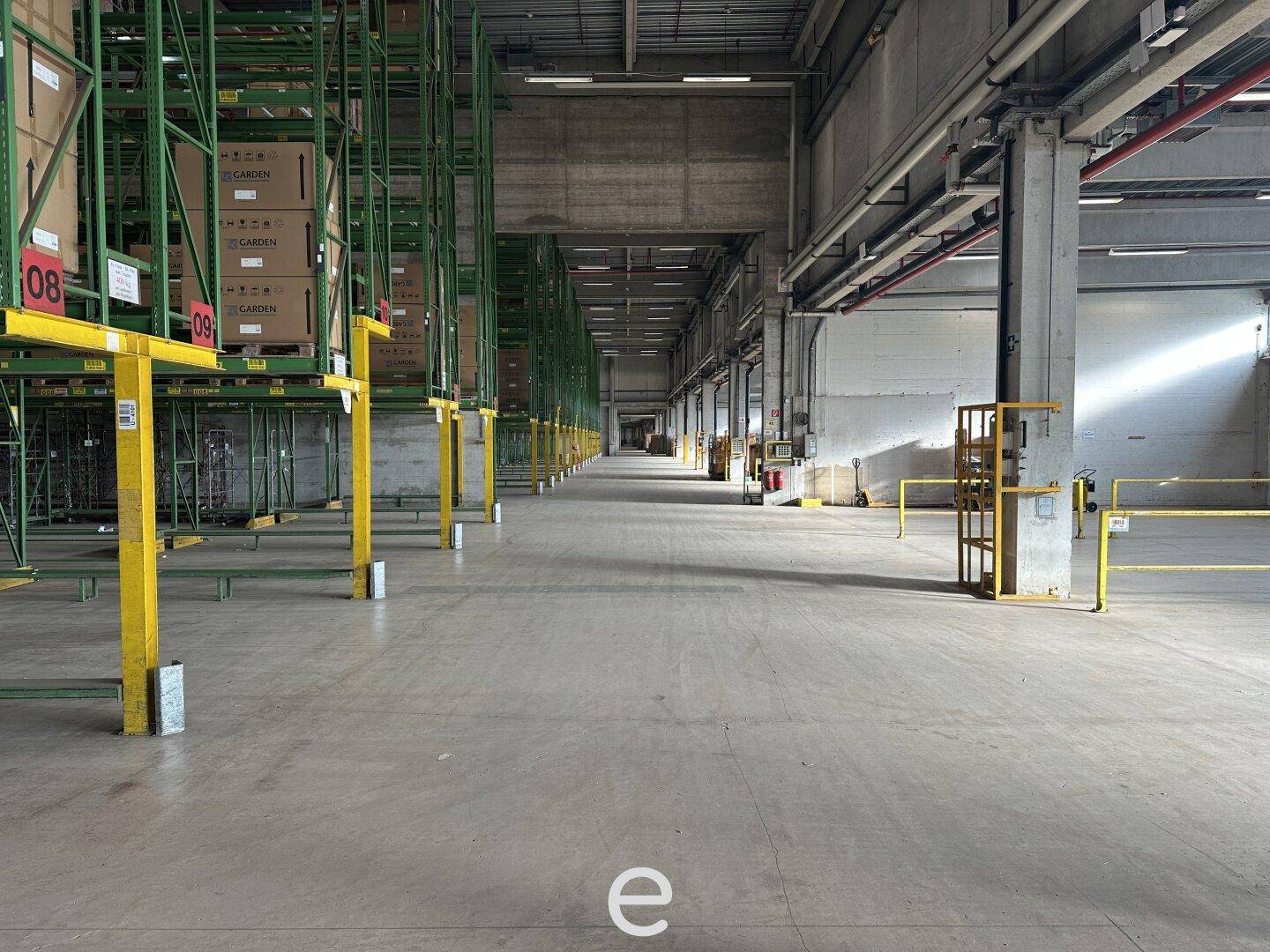 Leere Lagerhalle mit hohen Decken und Betonboden, teilweise mit grünen Regalsystemen ausgestattet.