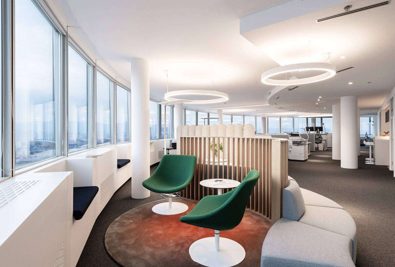 Gemütlicher Loungebereich im Büro mit grünen Sesseln, runden Teppichen und großen Fenstern mit Stadtblick.