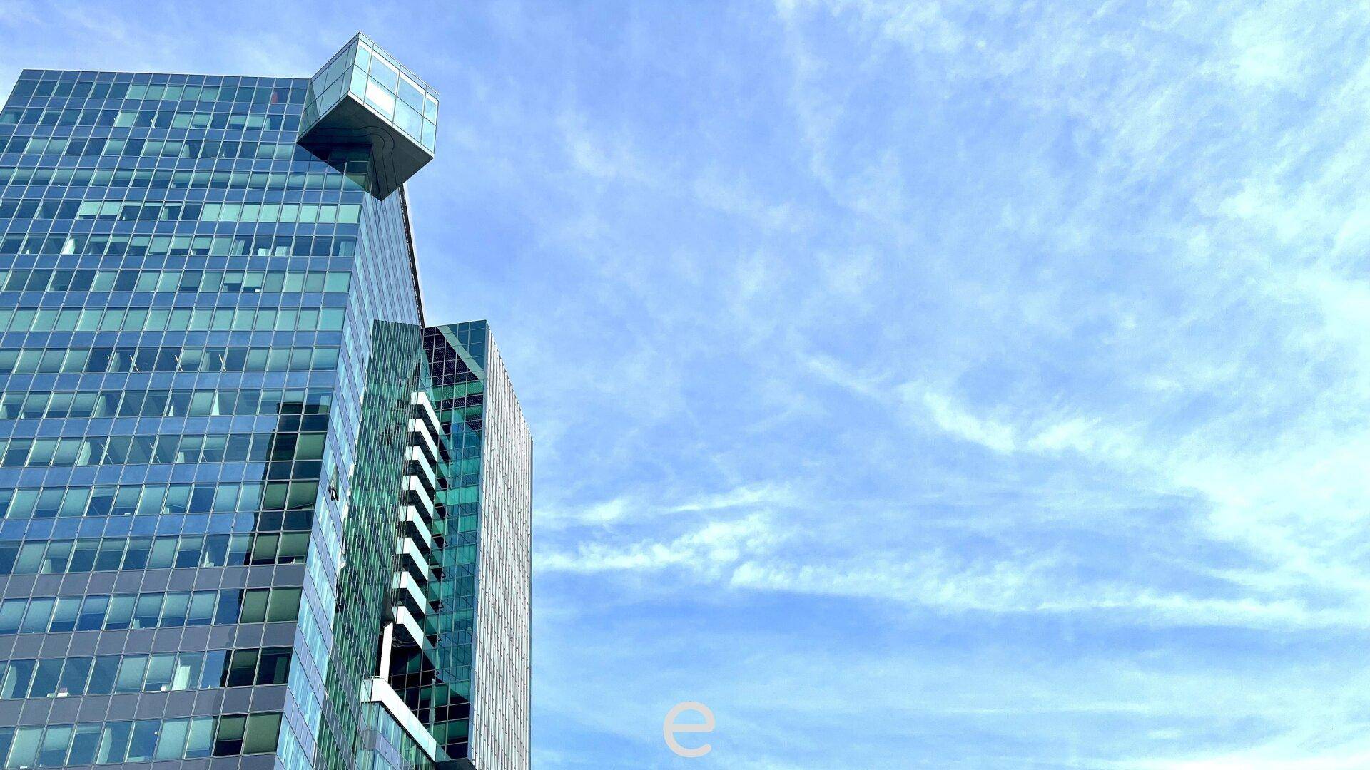 Detailansicht der oberen Etagen eines modernen Glasturms mit markanter Architektur vor blauem Himmel.